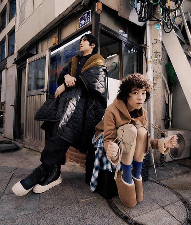 Two people pose on a city sidewalk by a building. One stands, leaning back in a quilted coat; the other crouches, wearing a tan jacket and slippers. Both look away thoughtfully. An address sign and stairway are visible above them.