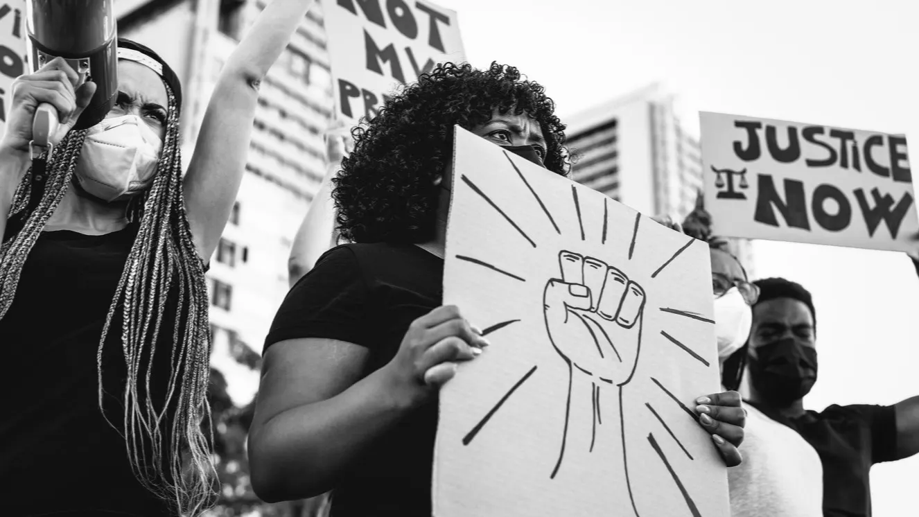 A group of protesters wearing masks hold signs, including one with a raised fist and another reading JUSTICE NOW, standing together in an urban setting with tall buildings in the background.