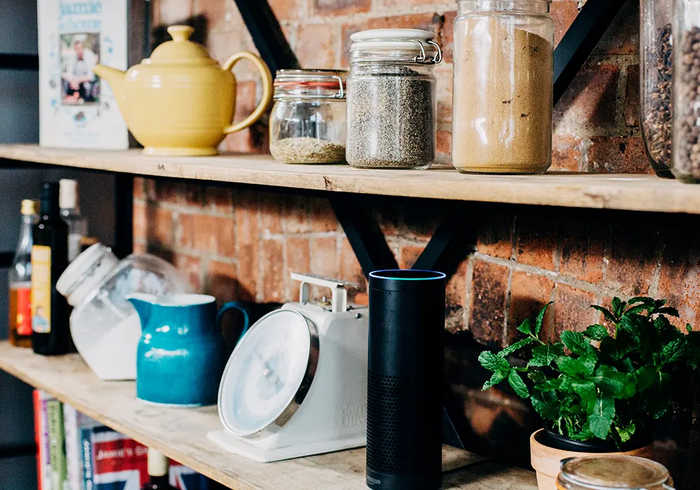 Wooden kitchen shelves with jars of spices, a teapot, a blue pitcher, a kitchen scale, an Amazon Echo speaker, a potted mint plant, and glass bottles, all against a brick wall background.
