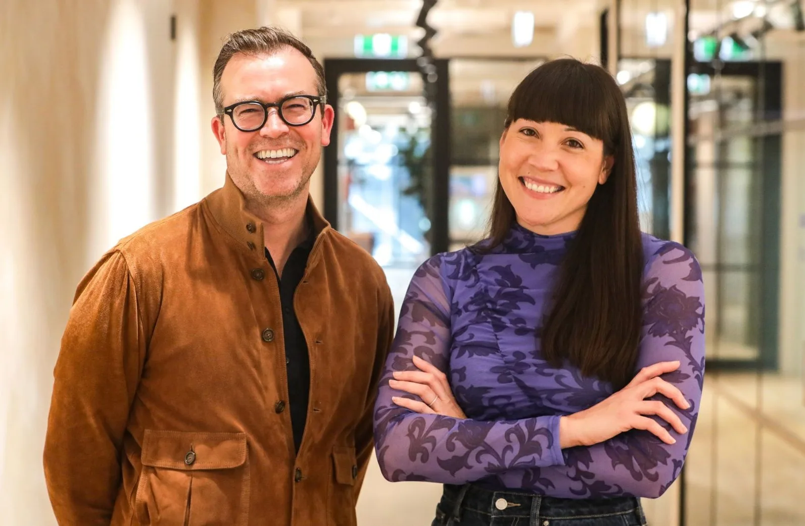 Two people stand indoors, both smiling. The man on the left wears glasses and a brown jacket. The woman on the right has long dark hair with bangs and wears a purple patterned top with arms crossed. The background shows a modern office hallway.
