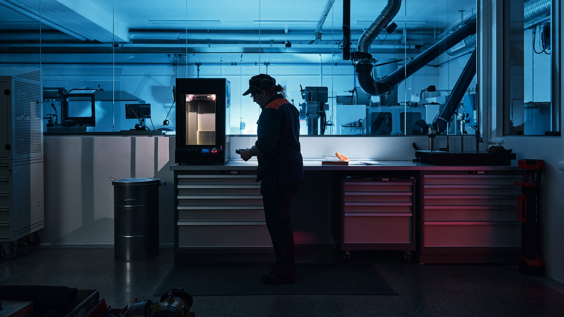 A person wearing a cap and safety glasses operates a machine in a modern industrial lab with blue lighting, surrounded by workbenches, drawers, and equipment.
