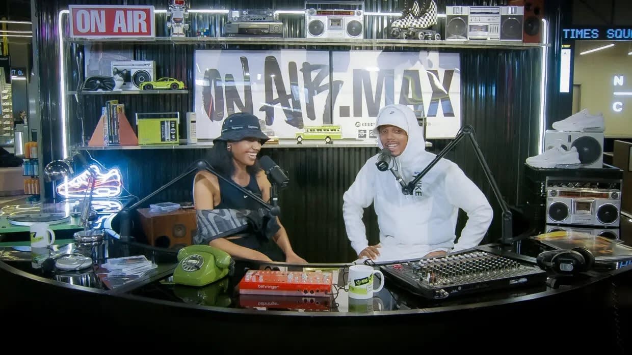 Two people sit at a radio station desk with microphones and audio equipment, smiling and talking. The background features a banner reading ON AIR: MAX, shelves with sneakers, and retro decor.