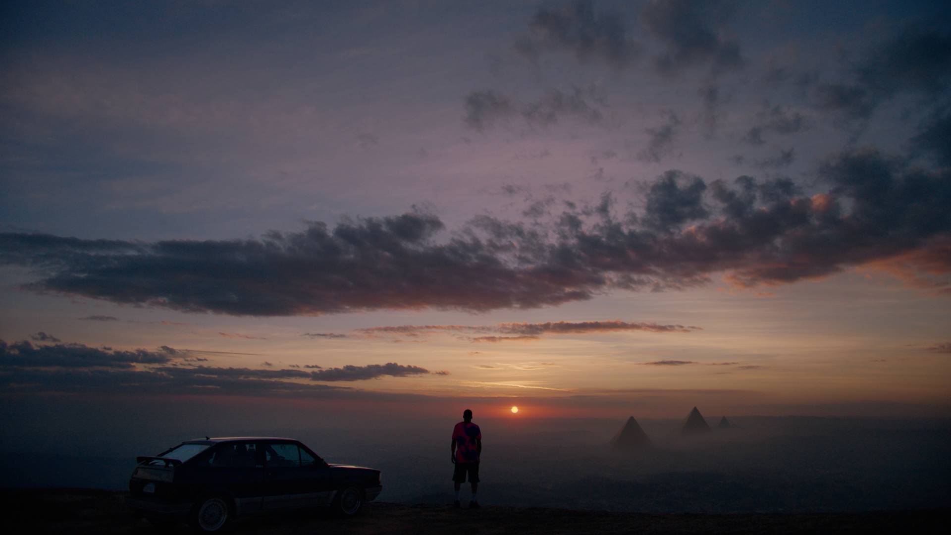A person stands next to a car on a hilltop at sunset, looking toward the horizon where the sun sets behind distant pyramids, with dramatic clouds filling the sky.