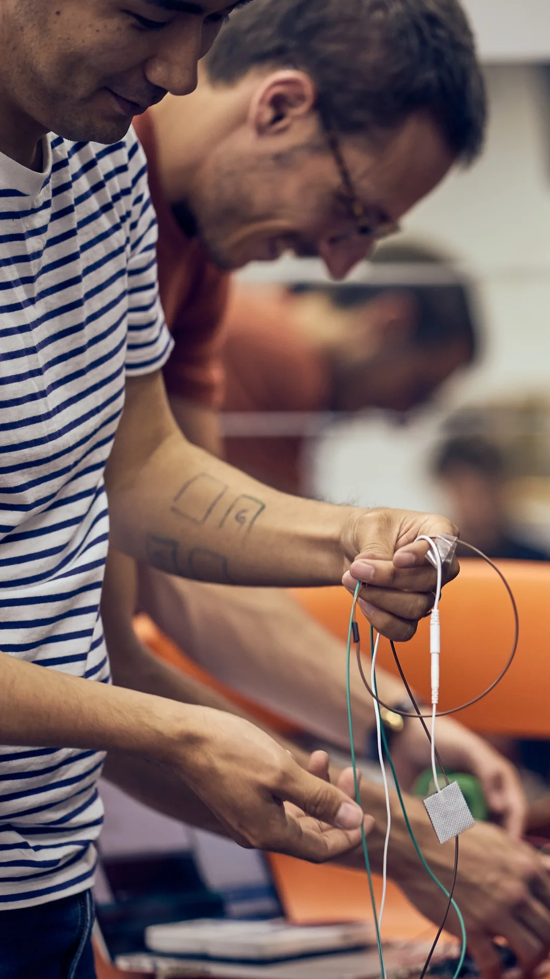 Two people work together with electronic components and wires. The person in the foreground wears a striped shirt and holds a device with several colored wires. Both seem focused on assembling or testing the equipment.