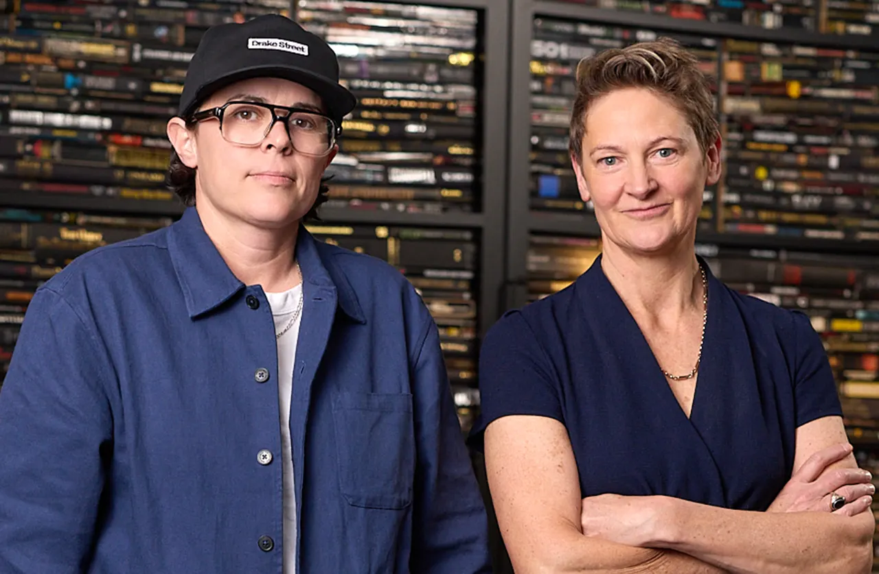 Two people stand in front of shelves filled with books. One wears glasses, a black cap, and a blue jacket; the other wears a navy dress and a gold necklace, with arms crossed. Both look at the camera and appear confident.