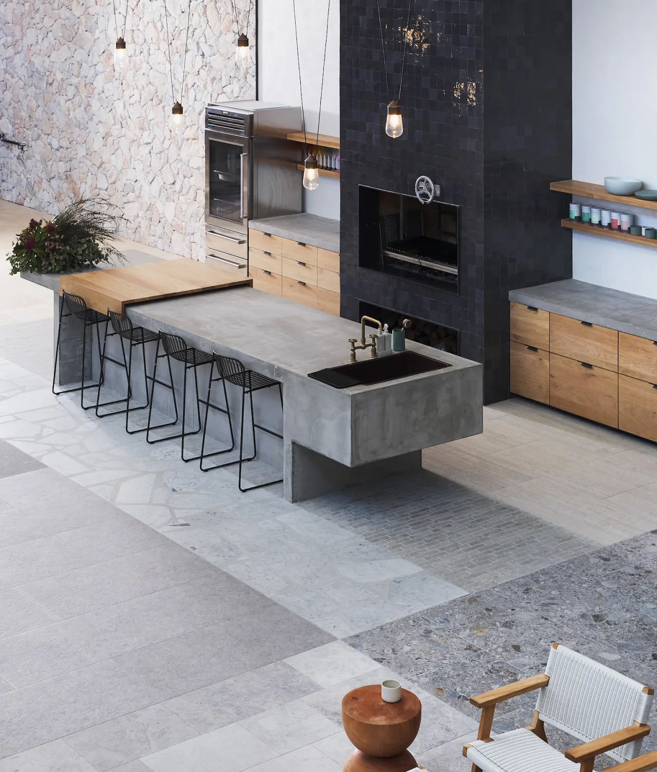Modern kitchen with a concrete island featuring a built-in sink, wooden cabinets, black tile backsplash, pendant lights, black wire bar stools, and a stone accent wall. A cozy seating area is visible in the foreground.