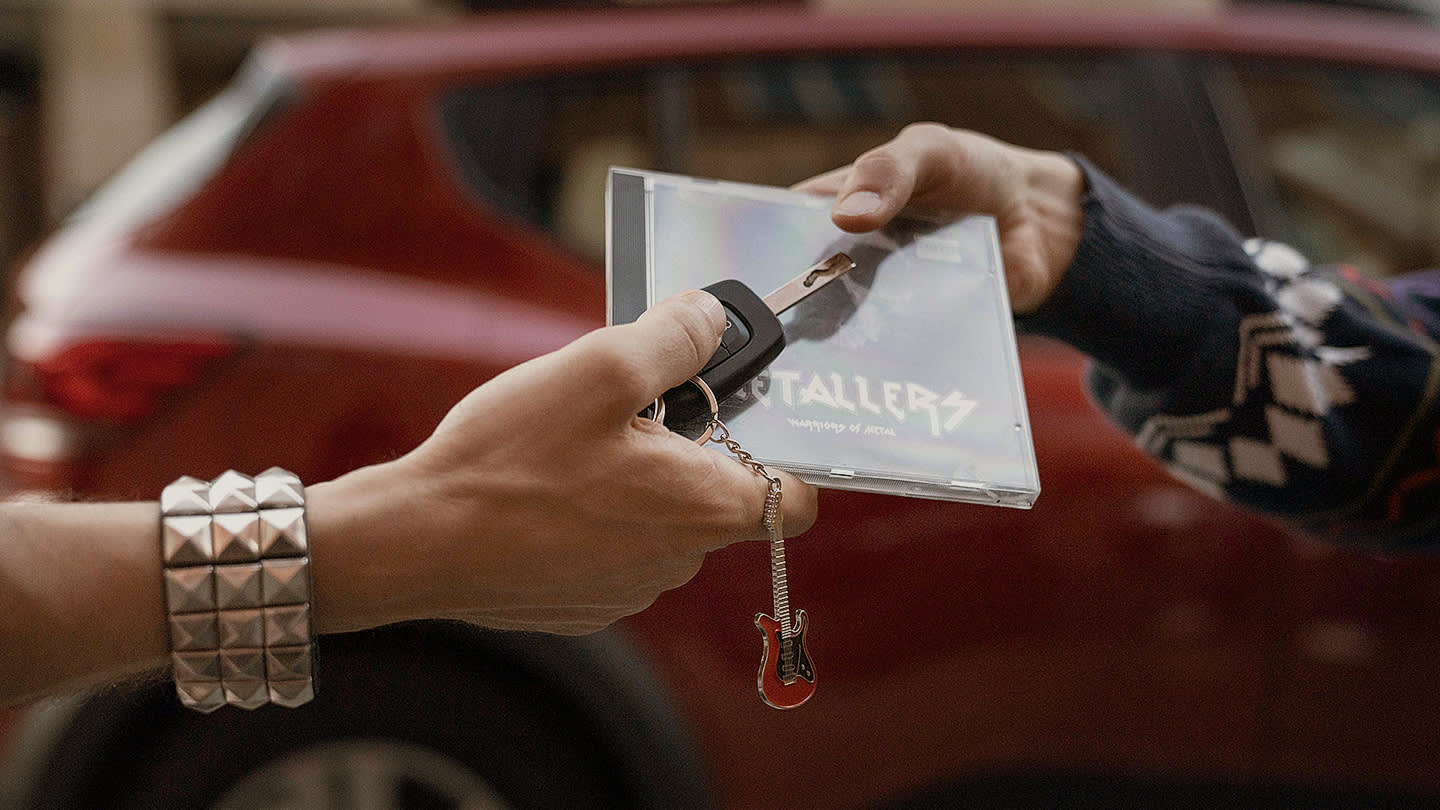 Two hands exchange a car key and a CD case in front of a red car. One hand wears a studded bracelet, while the other person wears a patterned sleeve.