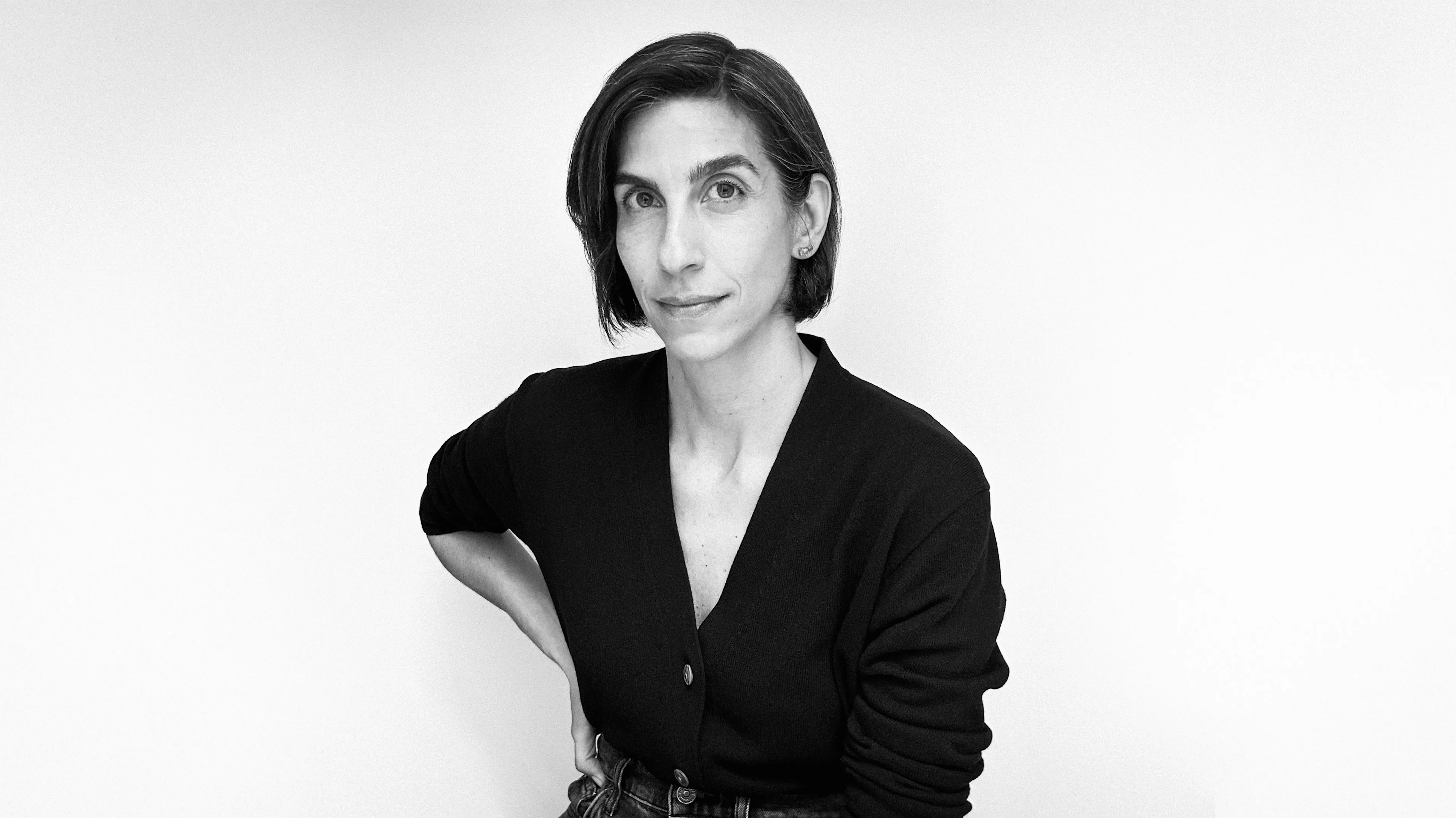 A person with short, dark hair and a black cardigan sits against a plain white background, looking directly at the camera with a neutral expression. One arm is bent and resting on their hip.