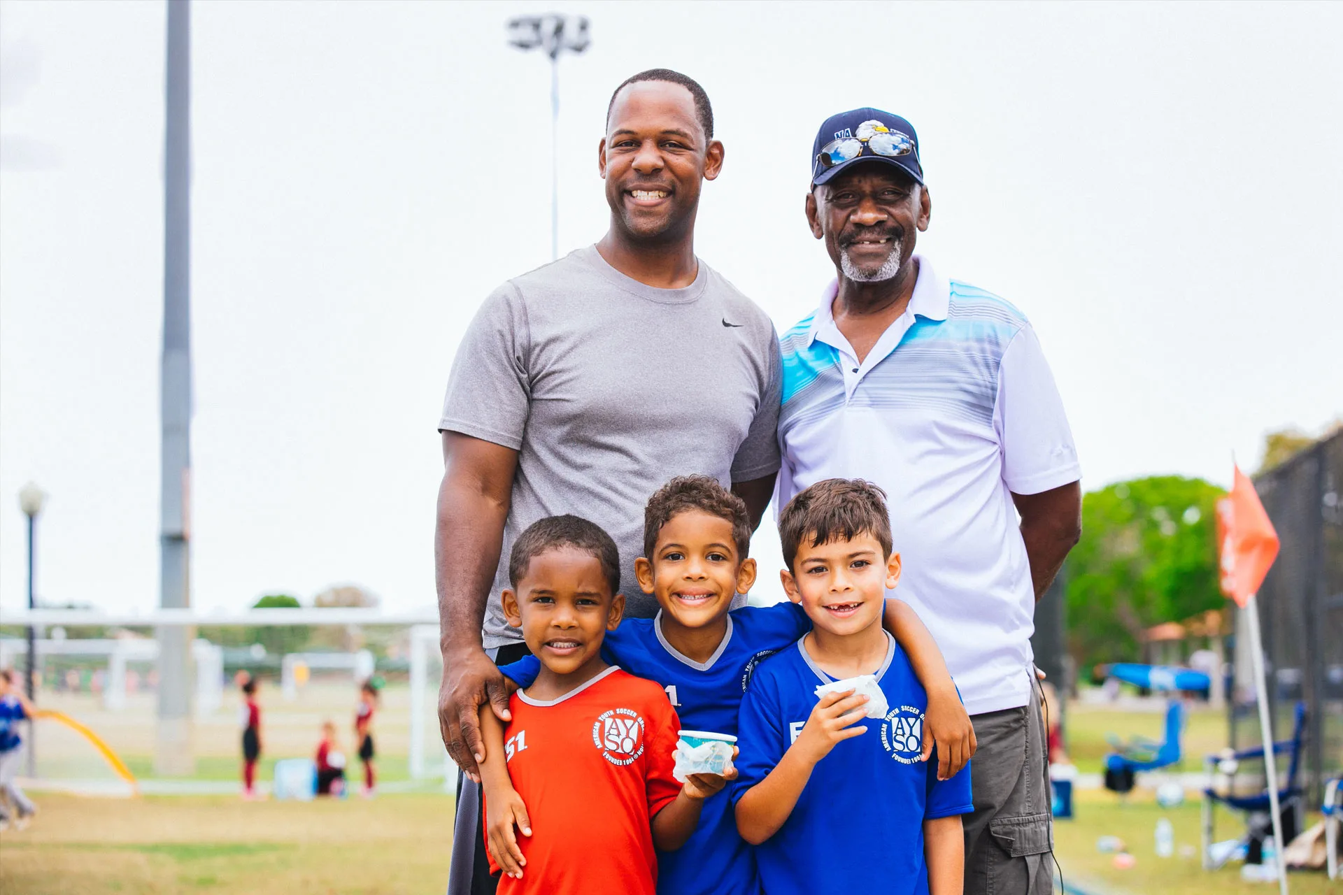 Two men stand smiling behind three young boys in soccer uniforms on a grassy field. The boys hold snacks and wear red and blue jerseys. Soccer goals and people are visible in the background.