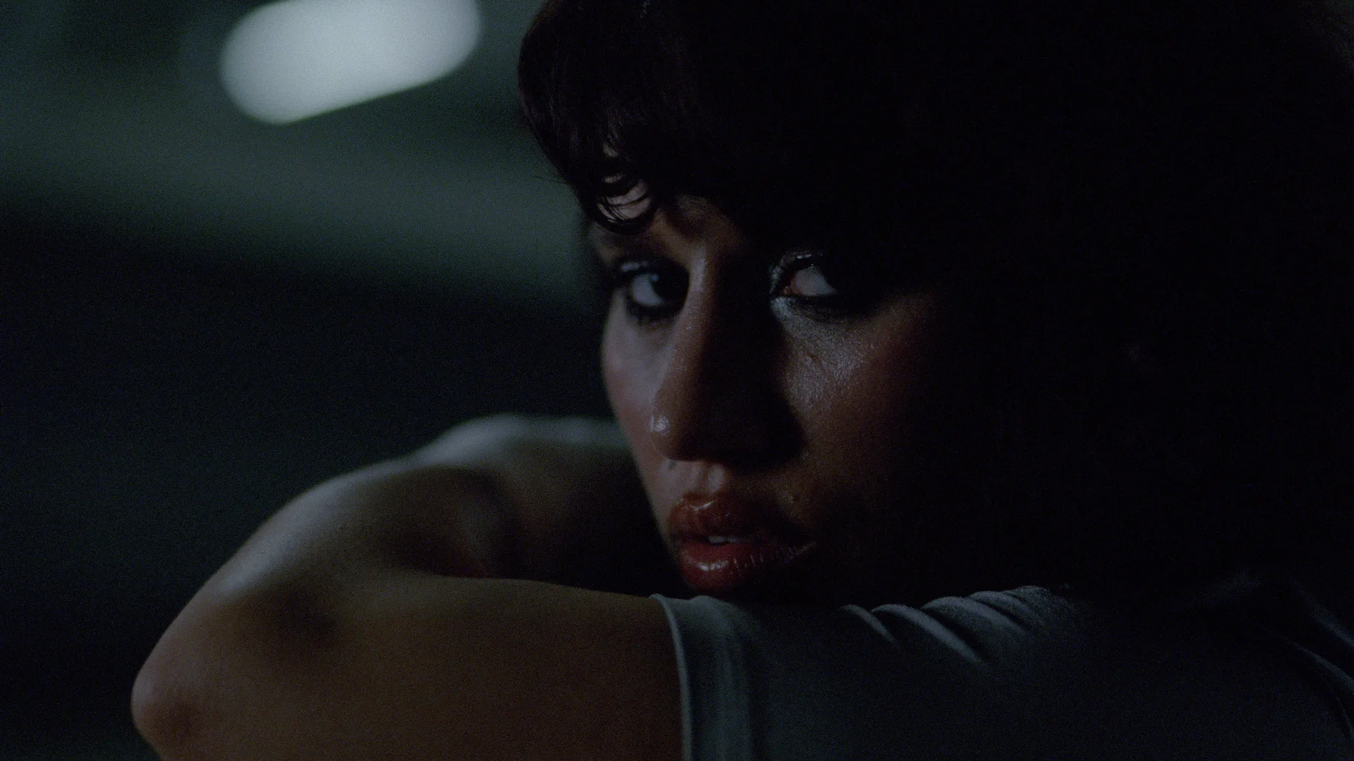 A person with dark hair and dramatic eye makeup looks over their shoulder in dim lighting, their face partially shadowed, creating a moody and intense atmosphere.