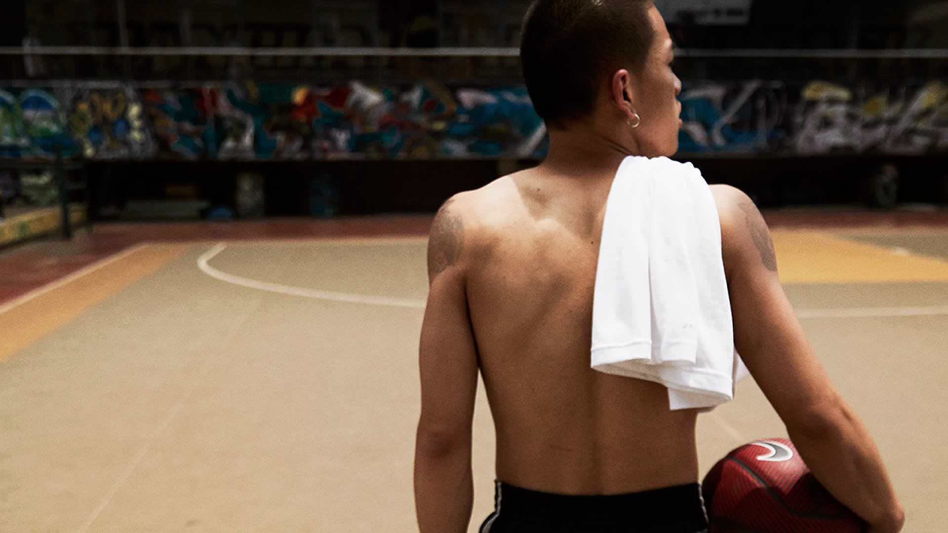 A shirtless person stands on an empty indoor basketball court, holding a basketball in one hand and a white shirt draped over their shoulder, facing away from the camera.