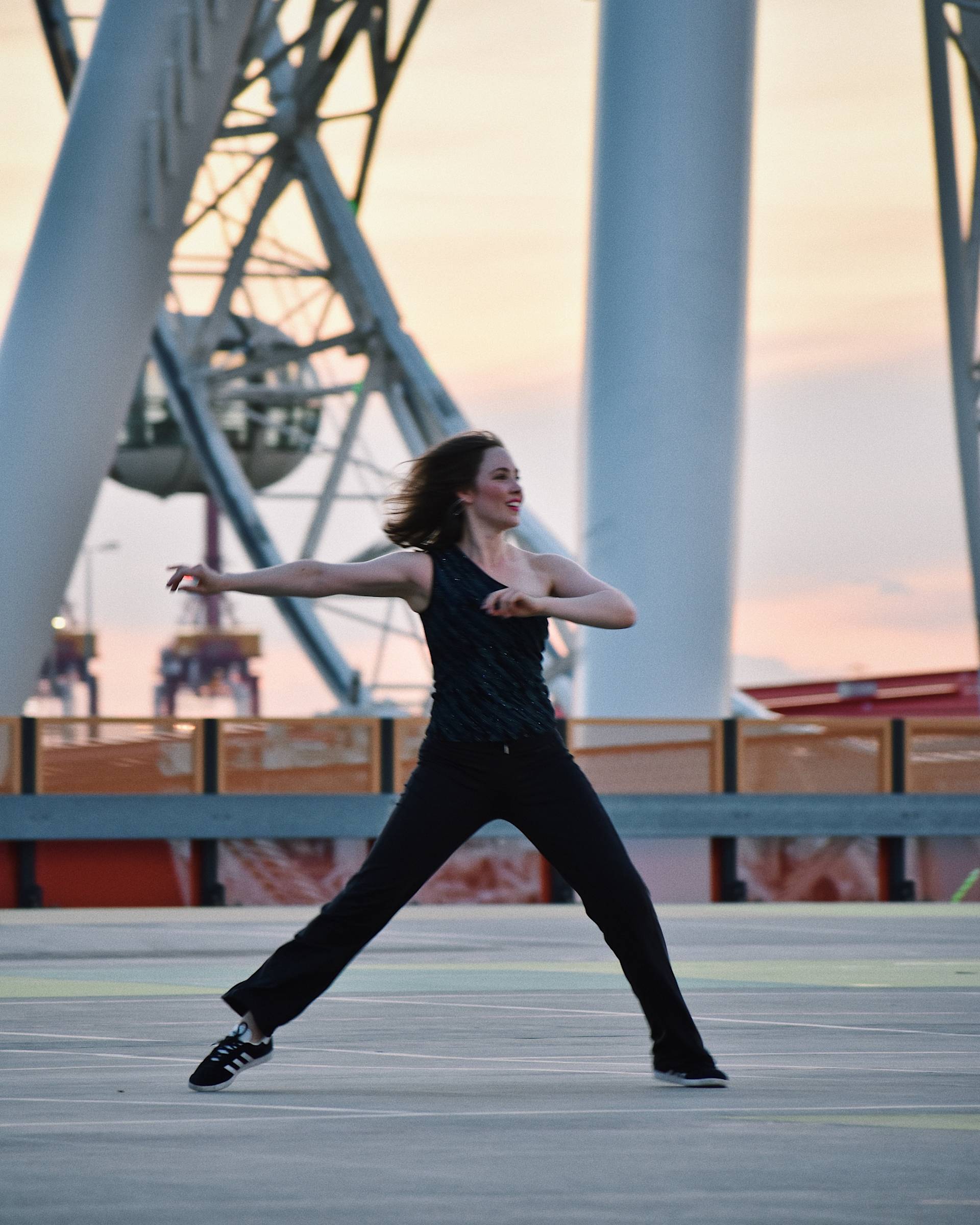 A woman wearing a black sleeveless top and black pants dances energetically outdoors at sunset, with large white structural beams and a ferris wheel in the background.
