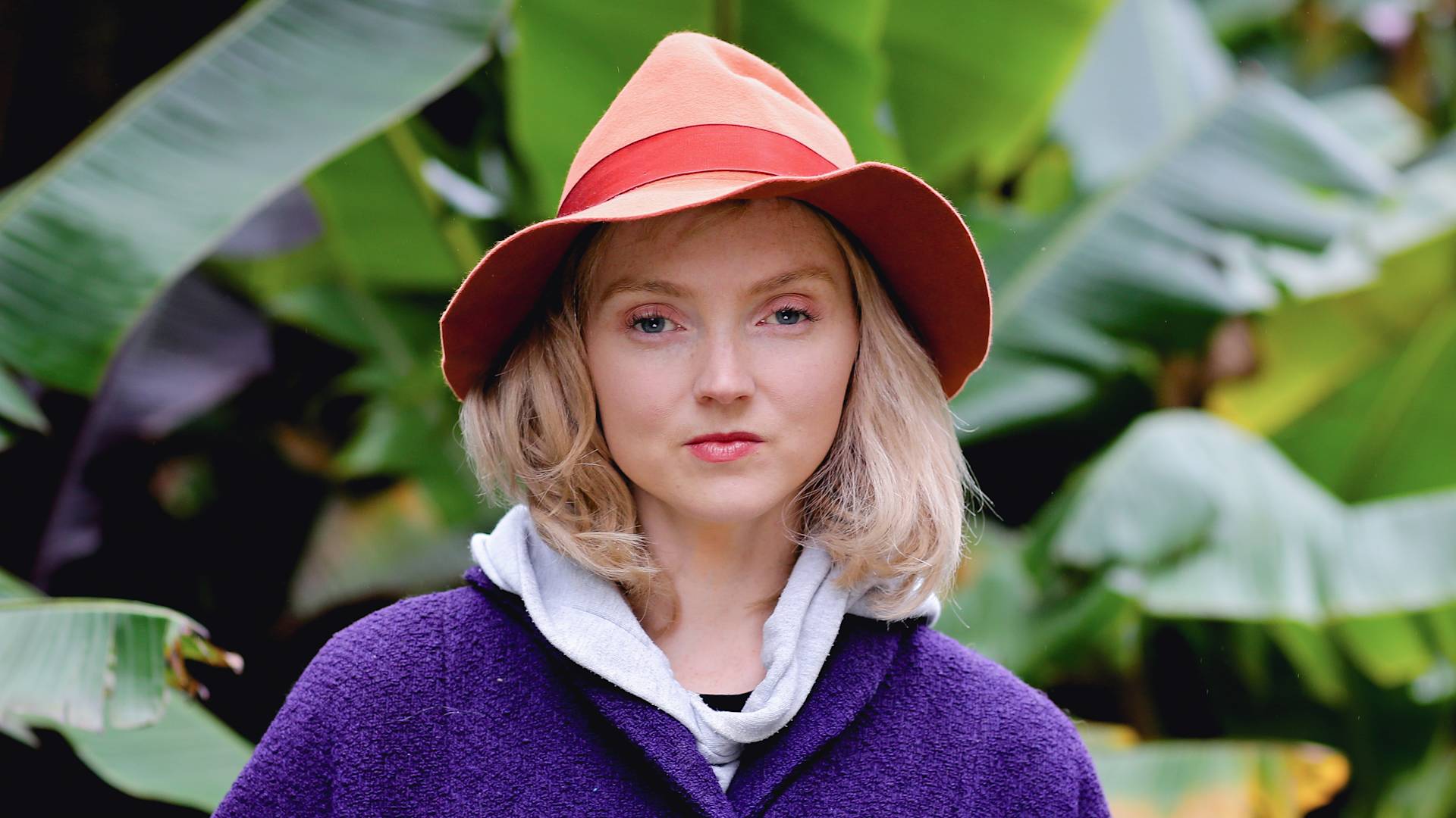 A woman with blond hair wearing an orange wide-brimmed hat, a gray hoodie, and a purple coat stands in front of large green tropical leaves, looking directly at the camera.