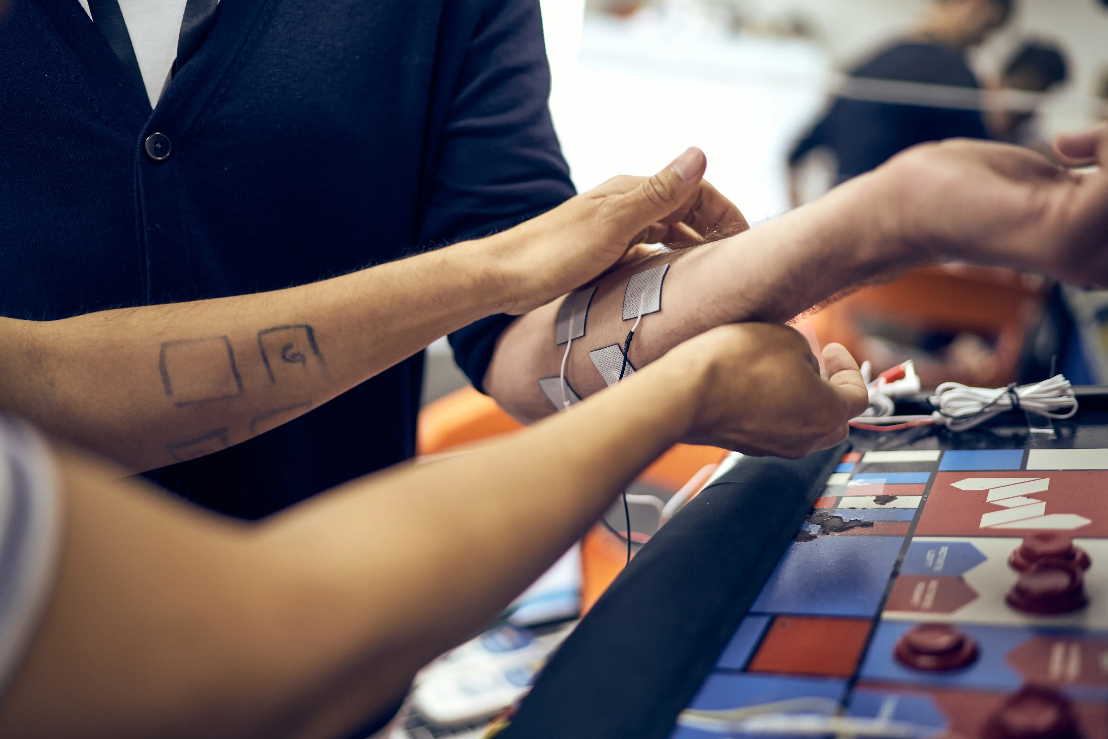 A person attaches small square electrodes to another persons forearm, possibly for a medical or physical therapy procedure, while standing beside a colorful game-like board on a table.