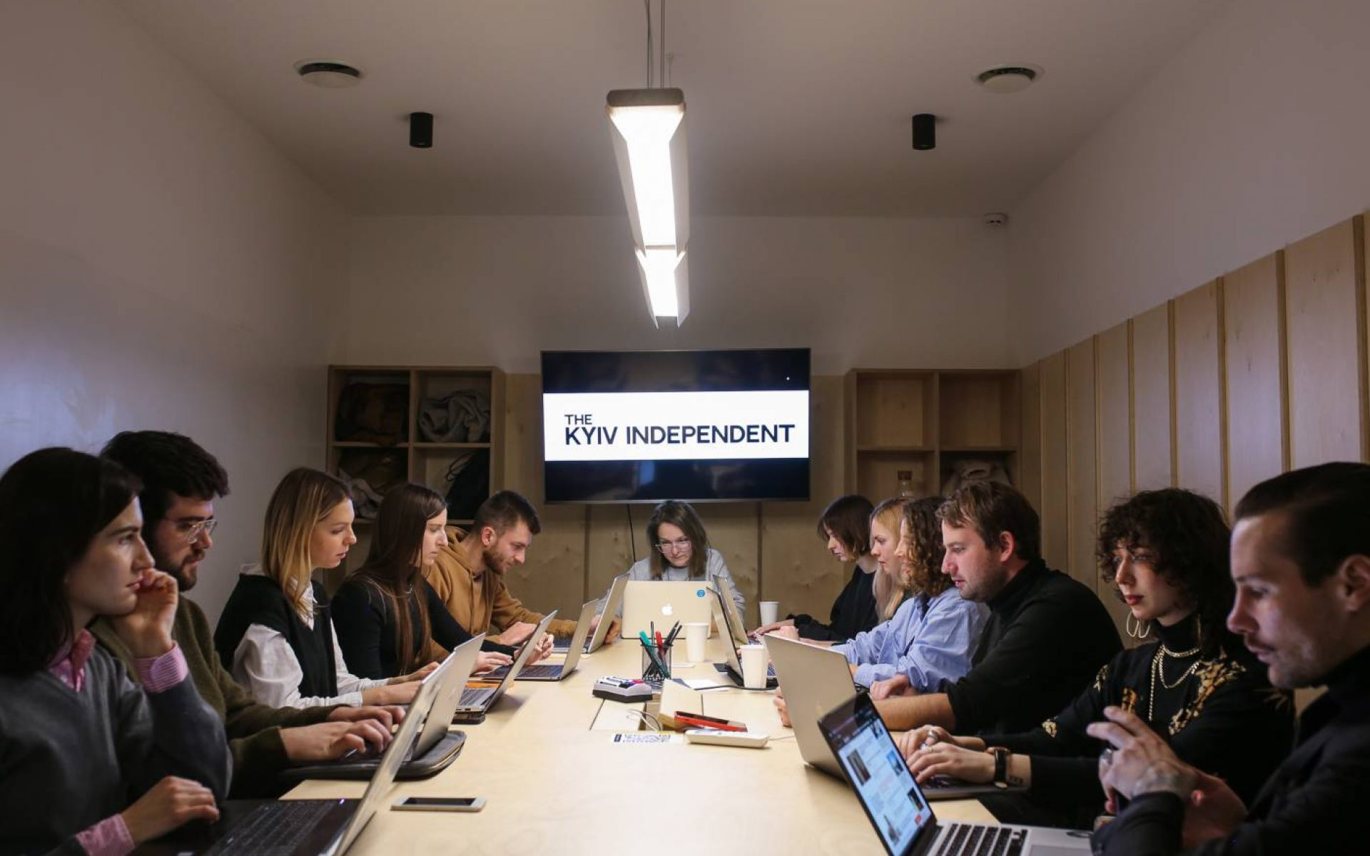 A group of people sit at a long conference table working on laptops, facing each other. A screen at the end of the table displays the words THE KYIV INDEPENDENT. The room has light walls and ceiling lights.