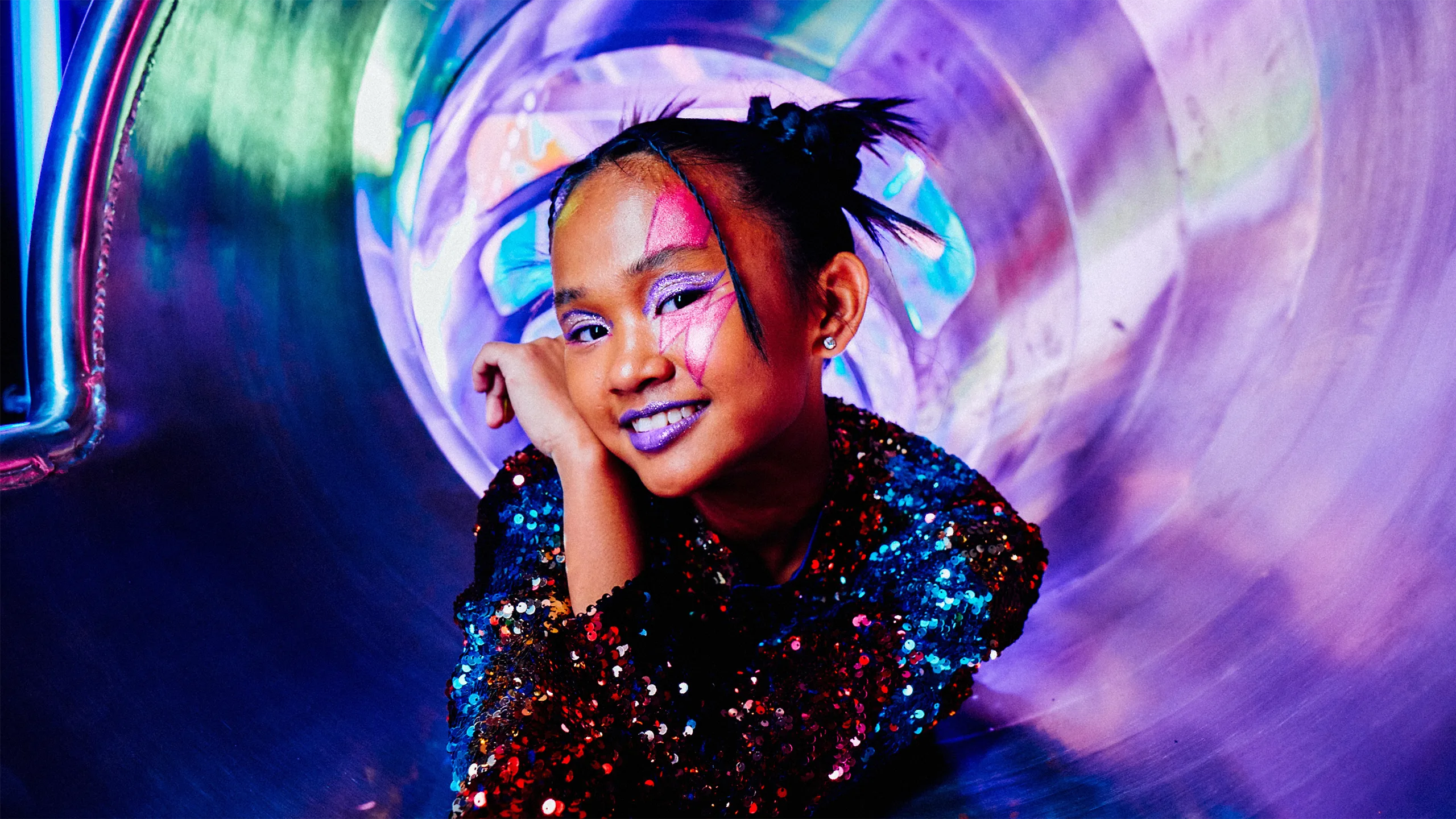 A smiling child with colorful face paint, sparkly makeup, and a sequined outfit poses inside a shiny, iridescent tunnel, surrounded by vibrant purple and blue lights.