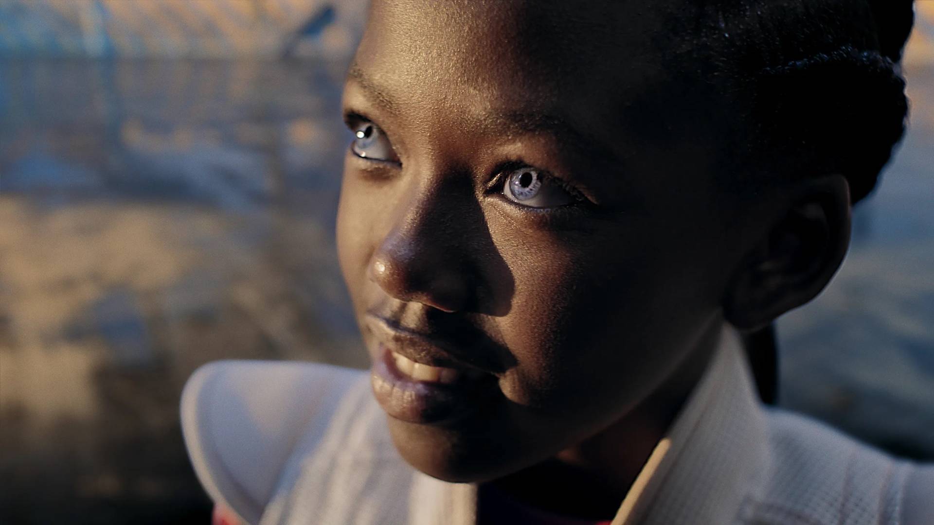 A close-up of a young person with dark skin and striking light blue eyes, looking upwards and smiling softly. The background is softly blurred with hints of sunlight and blue tones.