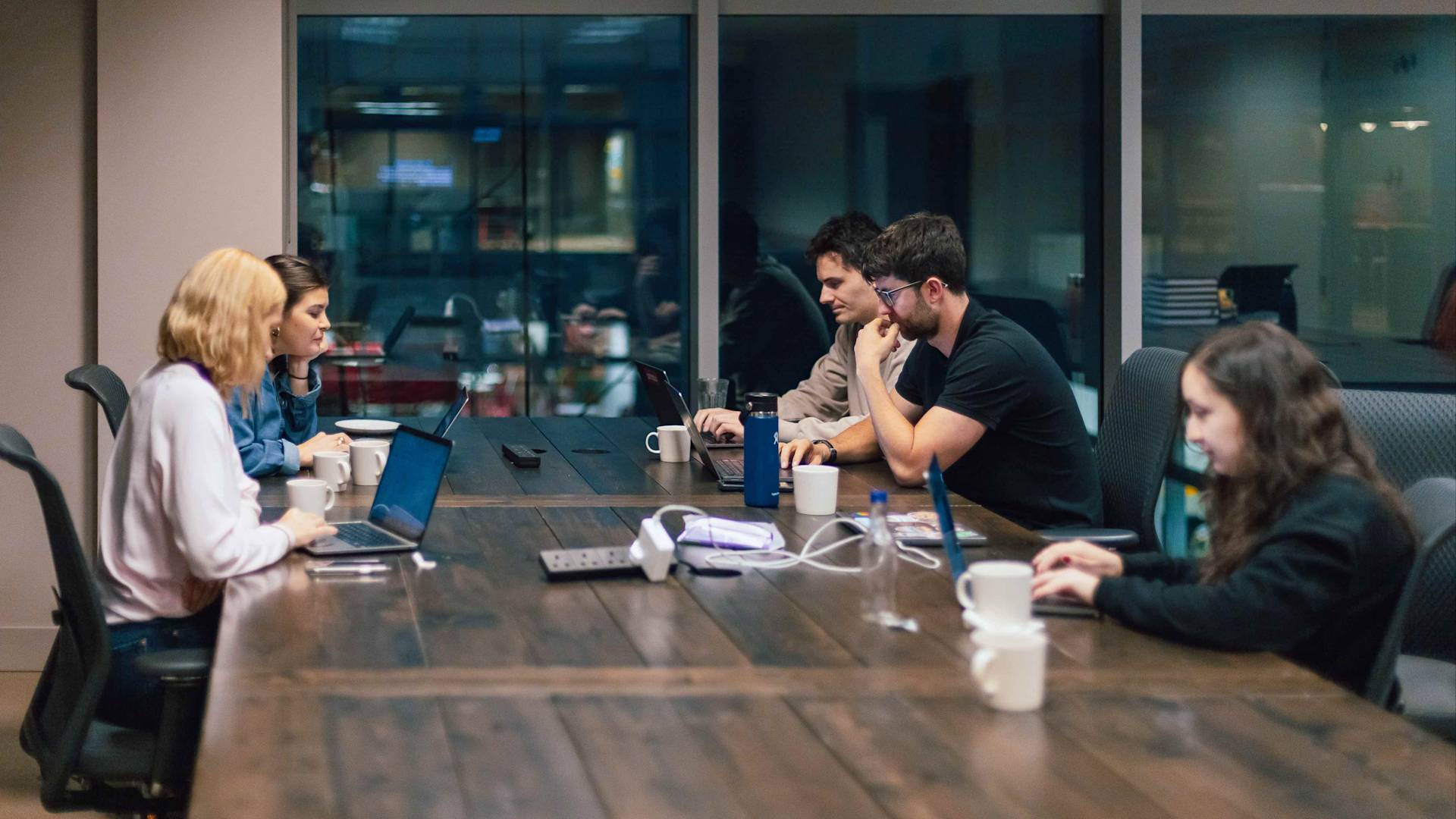 Six people sit around a large wooden conference table with laptops, mugs, and notebooks, engaged in discussion and work in a modern office meeting room with glass walls.