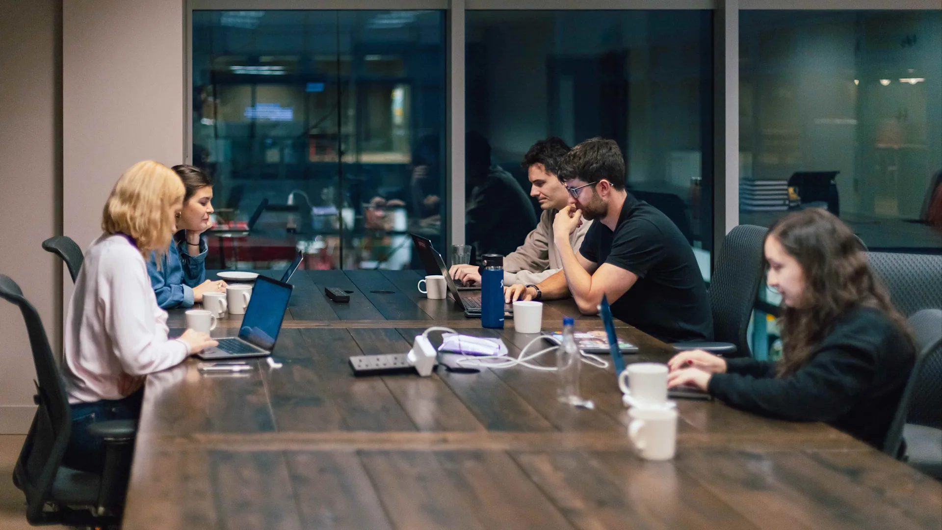 Six people sit around a large wooden conference table with laptops, mugs, and notebooks, engaged in discussion and work in a modern office meeting room with glass walls.