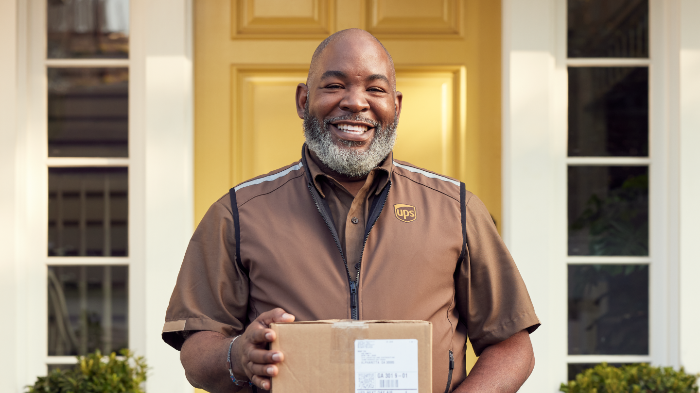 A smiling UPS delivery person stands in front of a yellow door, holding a package with a visible shipping label.