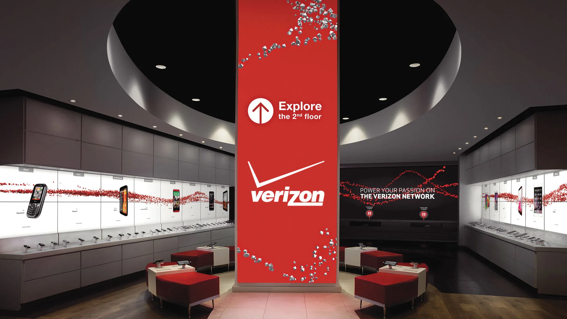 Modern Verizon store interior with mobile phones displayed along illuminated walls, a large red central pillar with the Verizon logo, and a sign directing customers to explore the second floor.