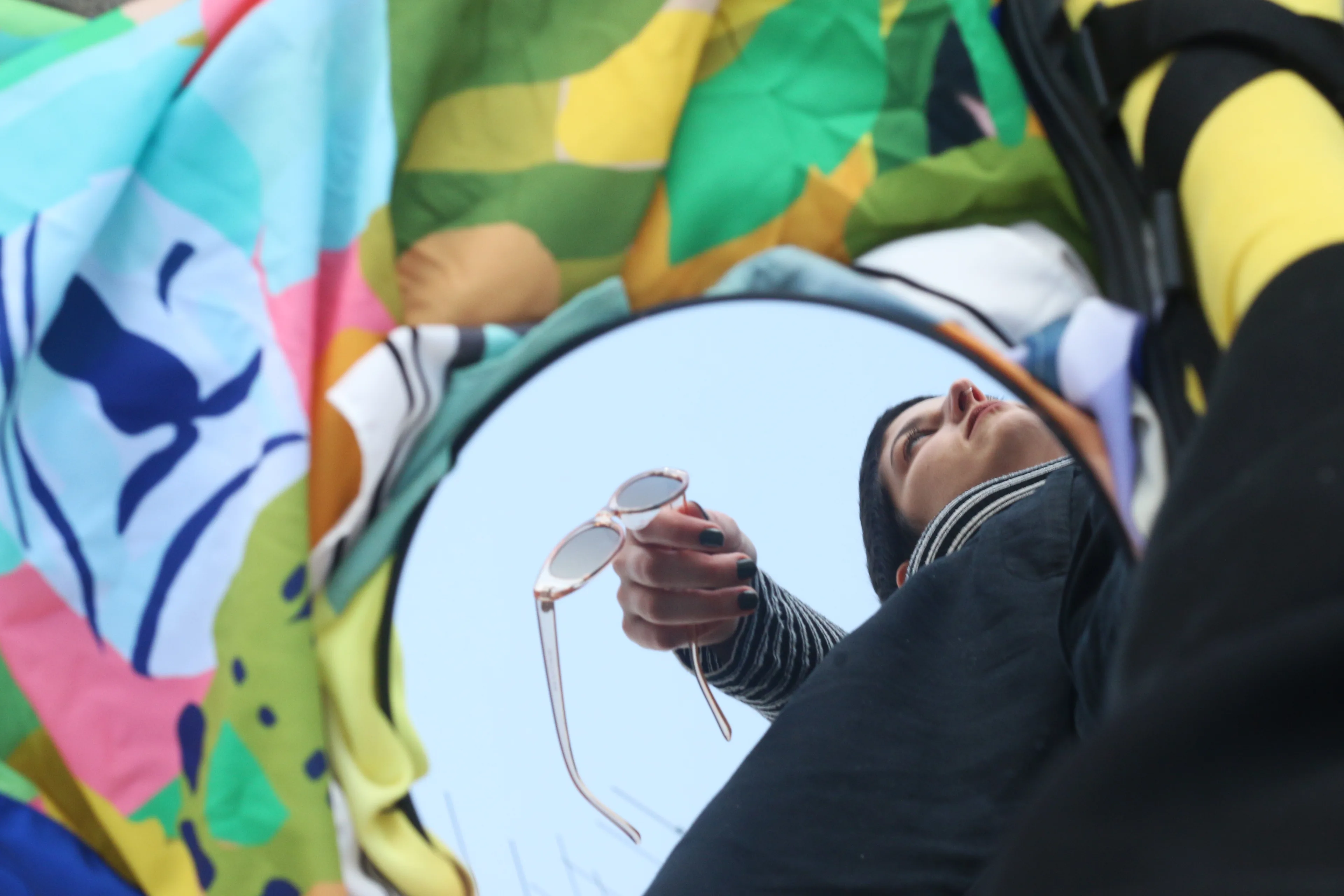 A person holding sunglasses is reflected in a round mirror, surrounded by colorful, patterned fabric. The image is taken from a low angle, creating an artistic and abstract composition.
