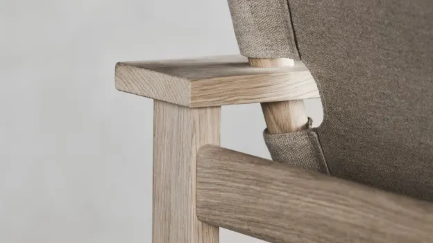 Close-up of the corner of a wooden chair with light-colored wood frame and beige fabric upholstery, showcasing the armrest and part of the backrest.