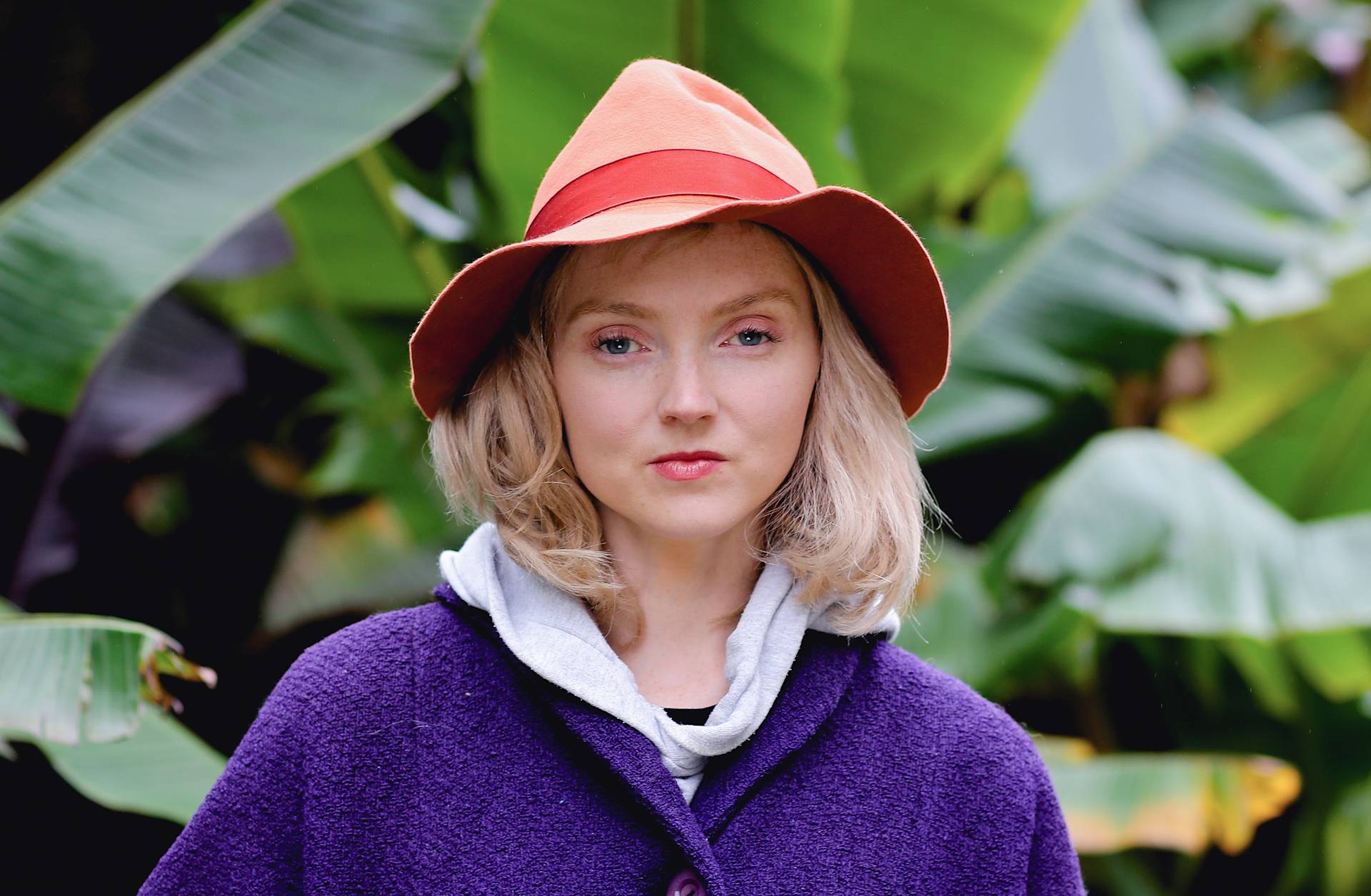 A woman with blonde hair wearing a two-tone orange hat, a purple coat, and a gray hoodie stands in front of large green banana leaves, looking directly at the camera.