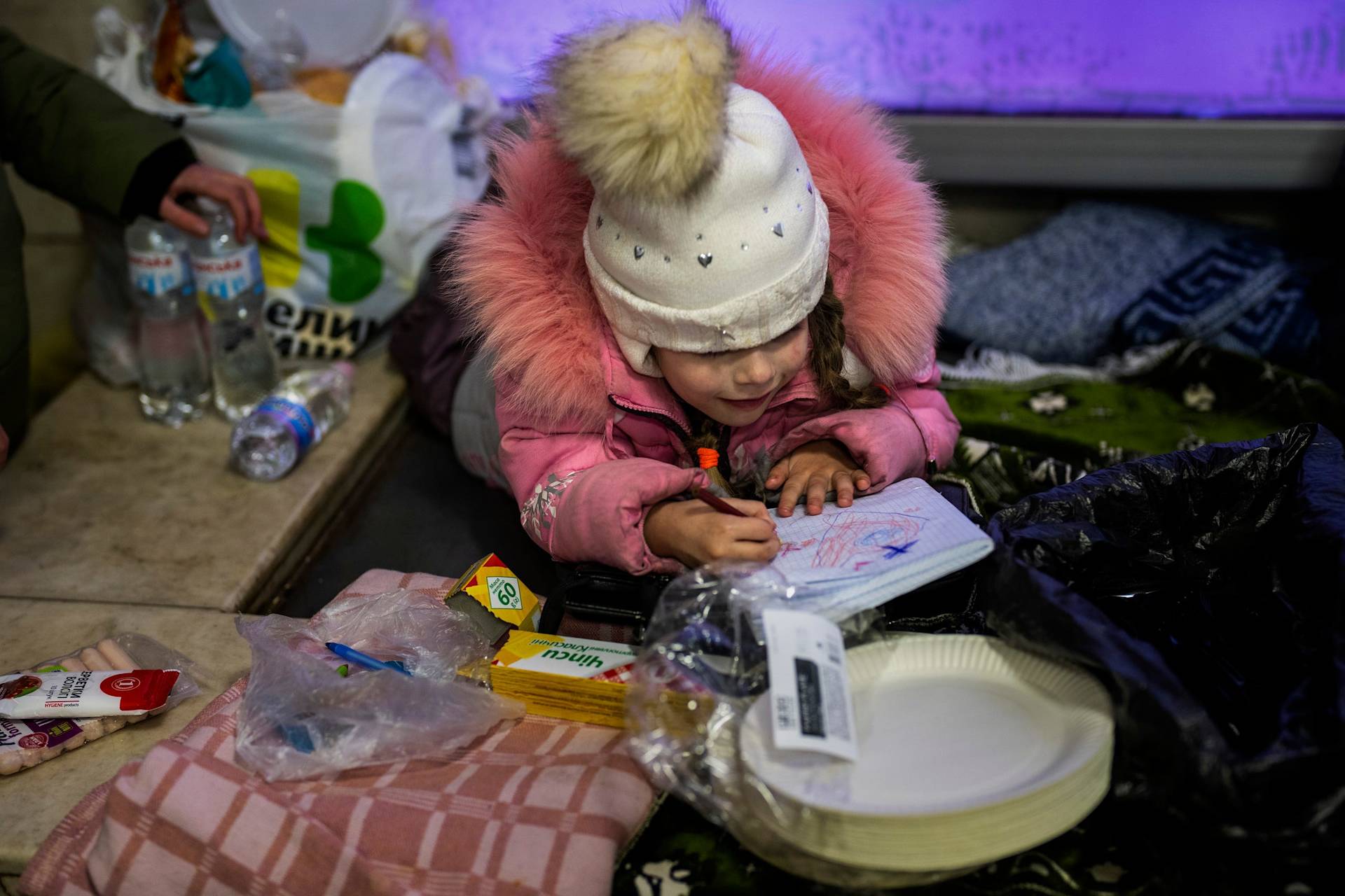 A young girl in a pink coat and white winter hat draws in a notebook while sitting among blankets and supplies, including water bottles, snacks, and paper plates.