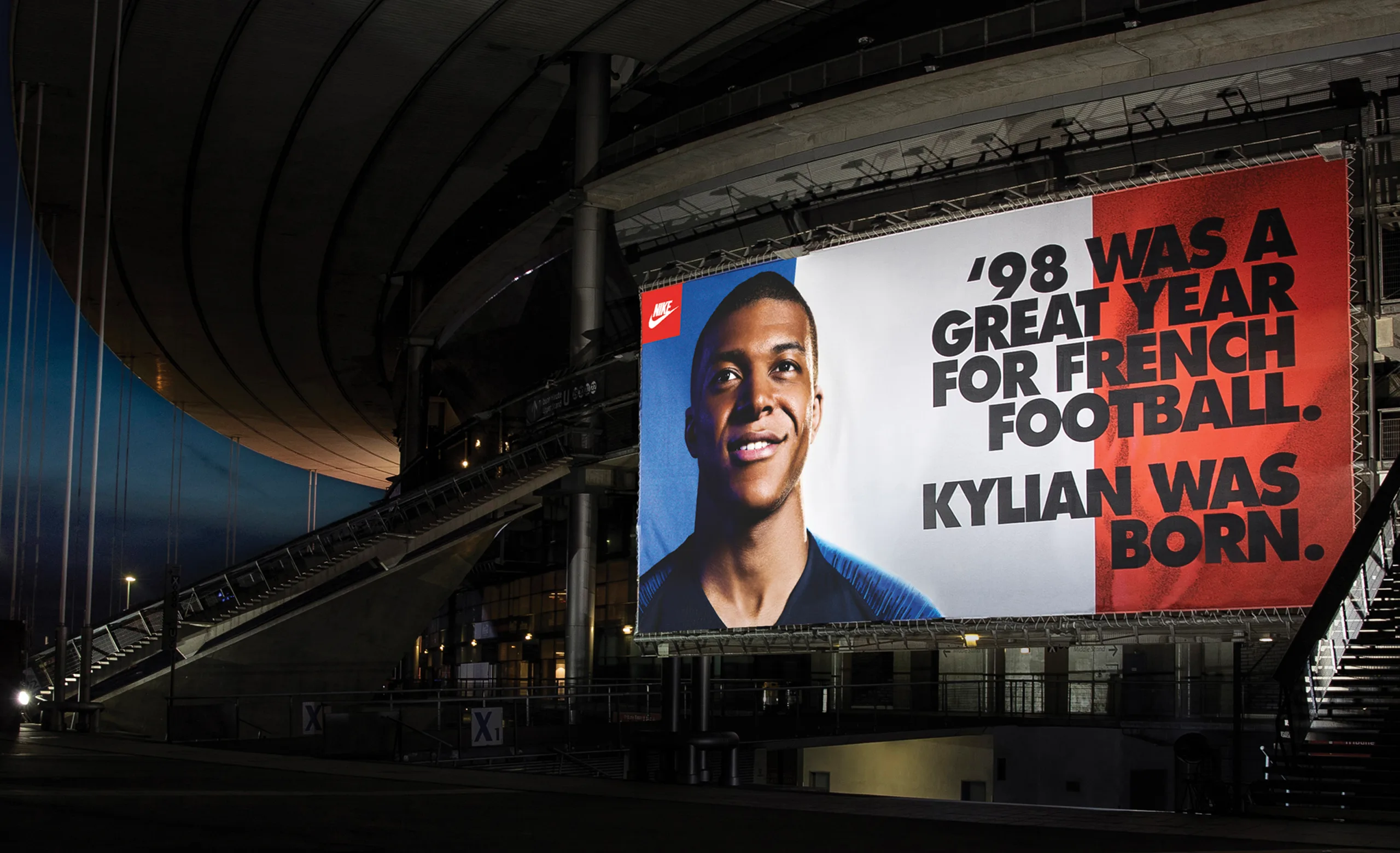 A large outdoor billboard features a smiling young man next to bold text: 98 was a great year for French football. Kylian was born. The billboard is displayed at a modern stadium at night.