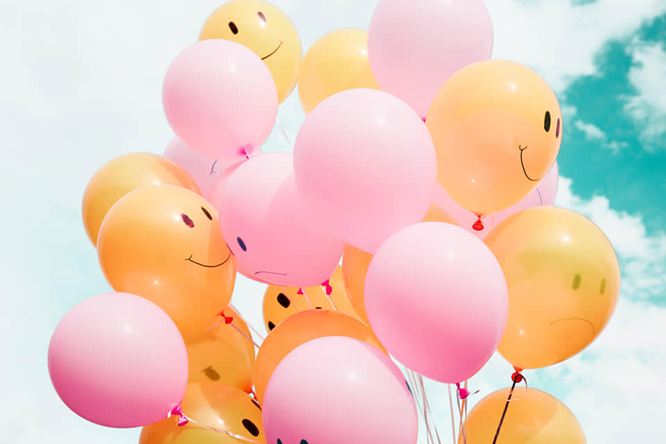 Cluster of pink and yellow balloons floating against a blue sky. The yellow balloons have happy or sad faces drawn on them, while the pink balloons are plain.