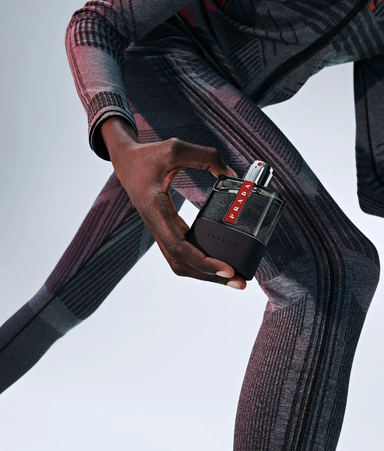 A person in a patterned, form-fitting gray outfit holds a bottle of Prada Luna Rossa Carbon perfume, positioned upright, against a light background. The focus is on the hand and the fragrance bottle.