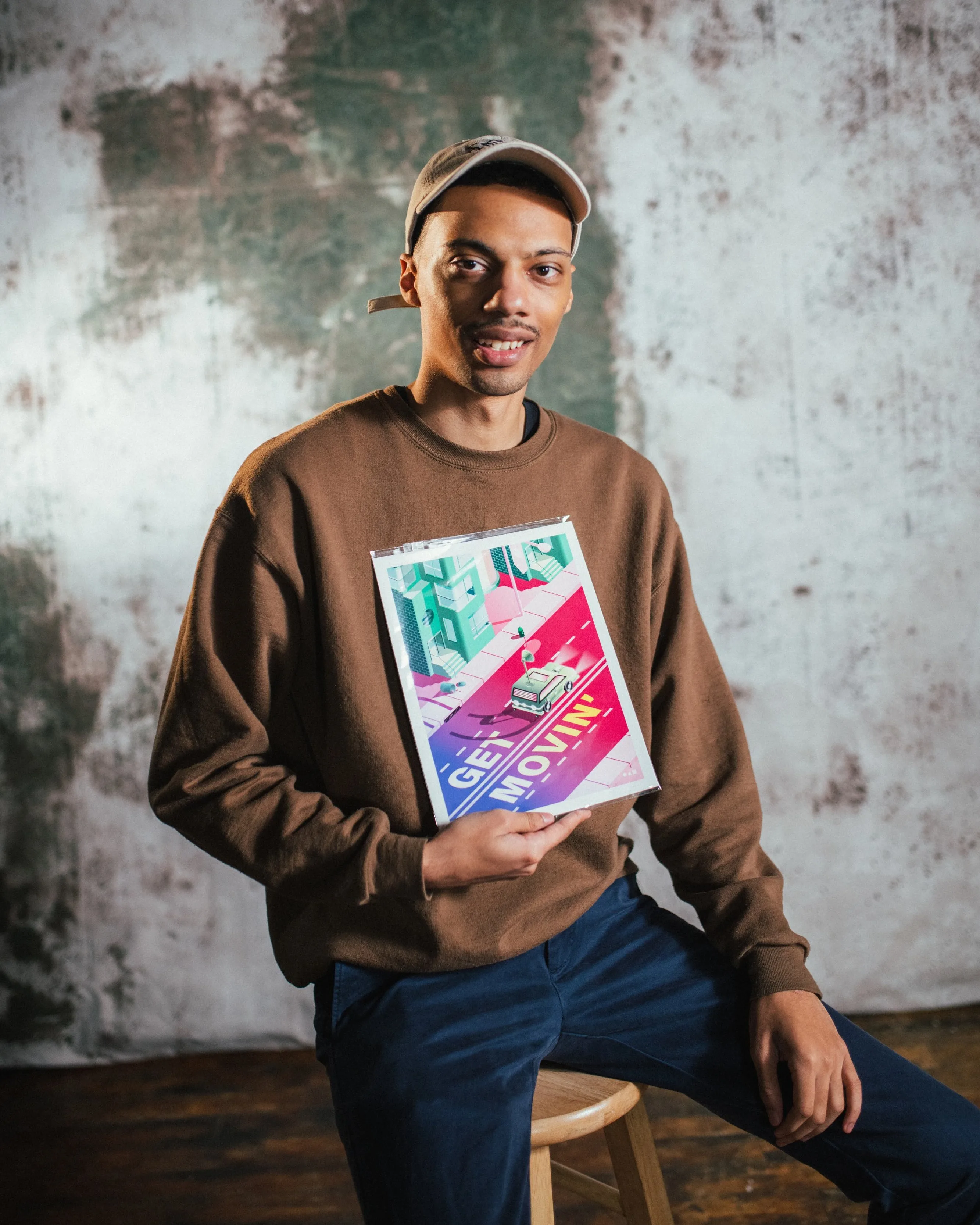 A young man wearing a brown sweatshirt, blue pants, and a beige cap sits on a stool, smiling and holding a colorful poster with the words GET MOVIN! in bold letters. The background is textured and mottled.
