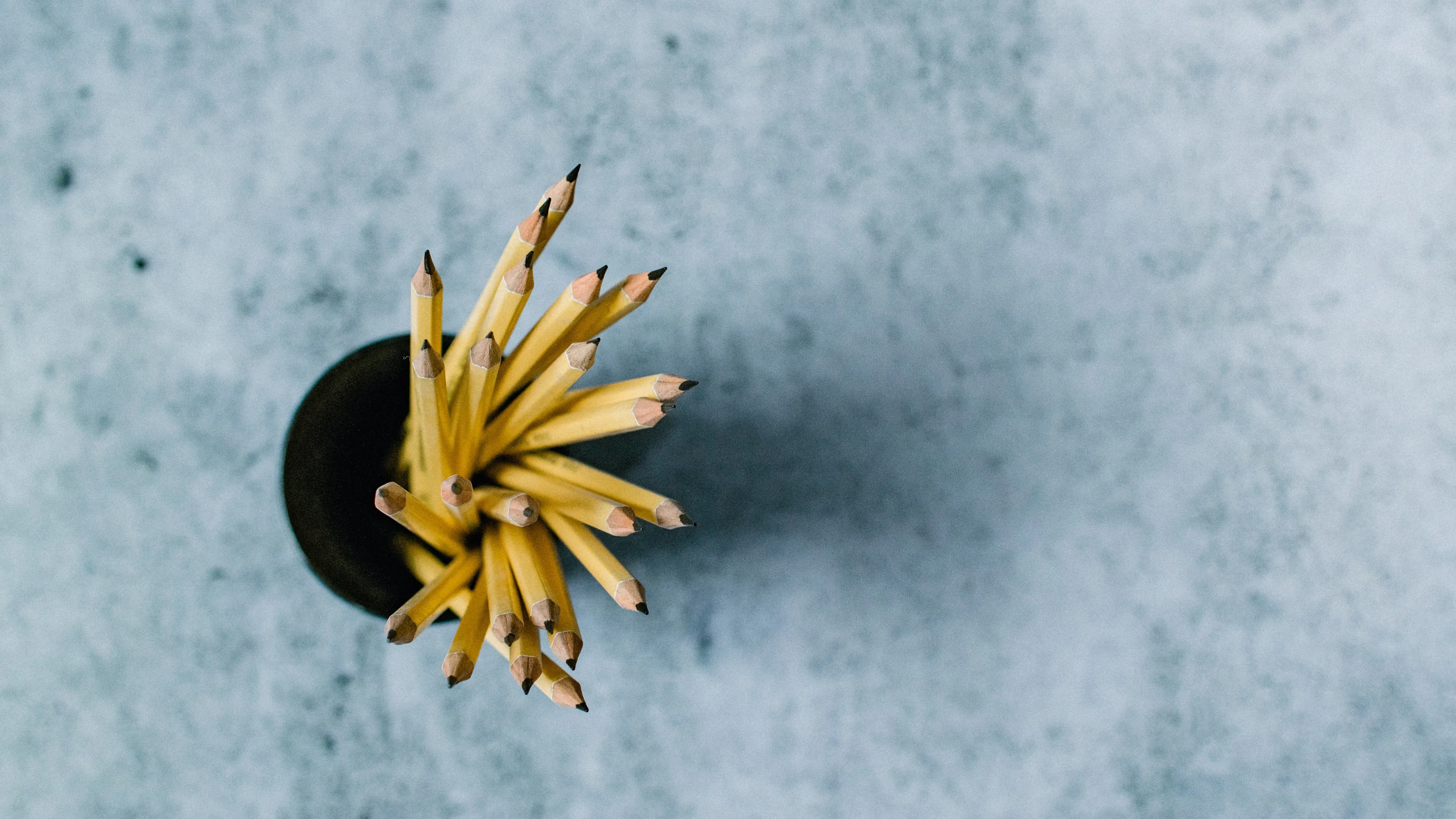 A top-down view of a black cup filled with sharpened yellow pencils, placed on a textured gray surface. The pencils are fanned out, and their pointed tips are visible.