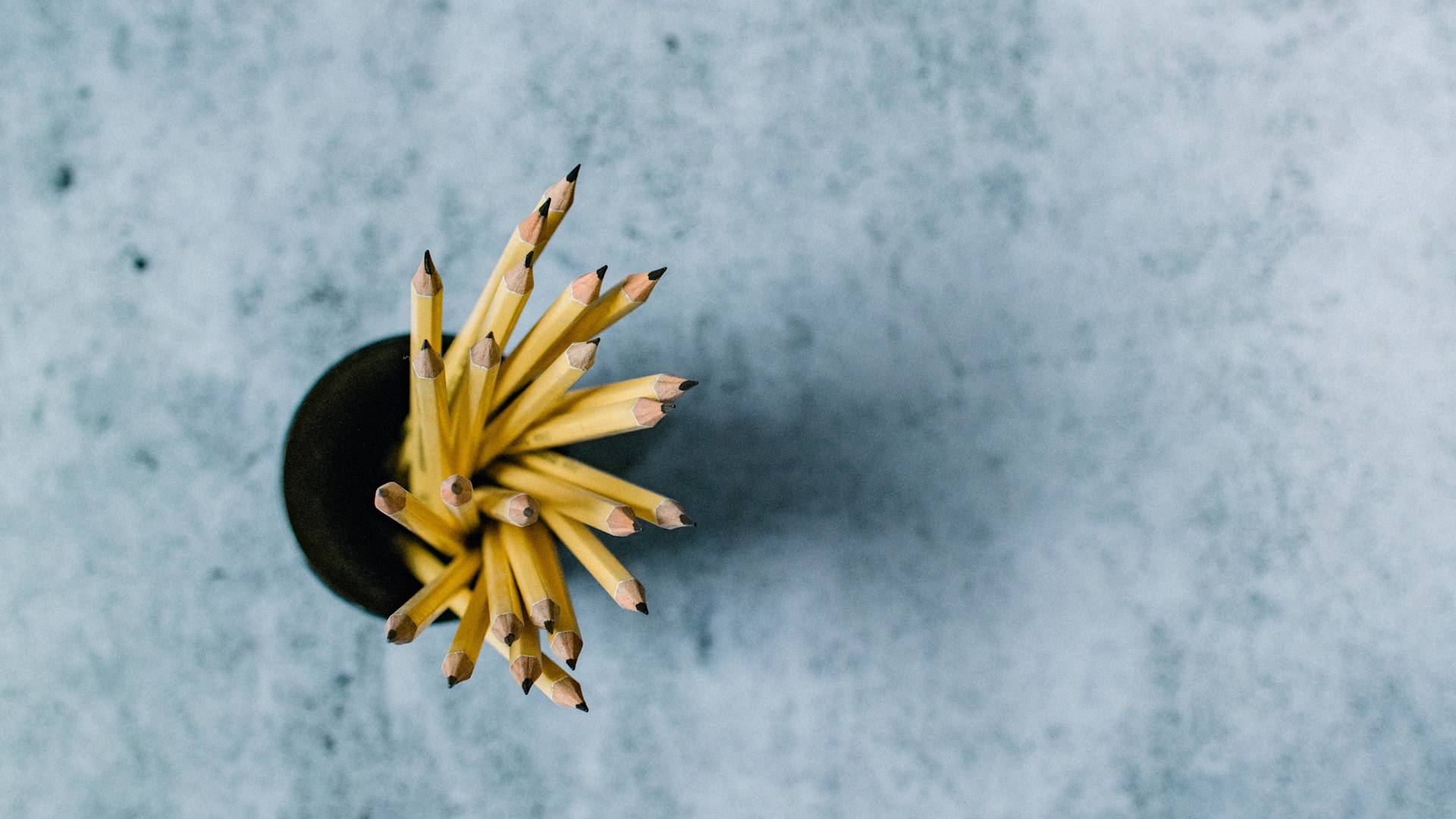 A top-down view of a black cup filled with sharpened yellow pencils, placed on a textured gray surface. The pencils are fanned out, and their pointed tips are visible.