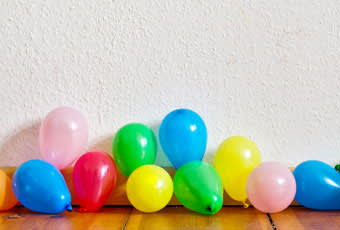 Colorful balloons in various shades of pink, red, blue, green, and yellow are scattered on a wooden floor against a plain white wall.