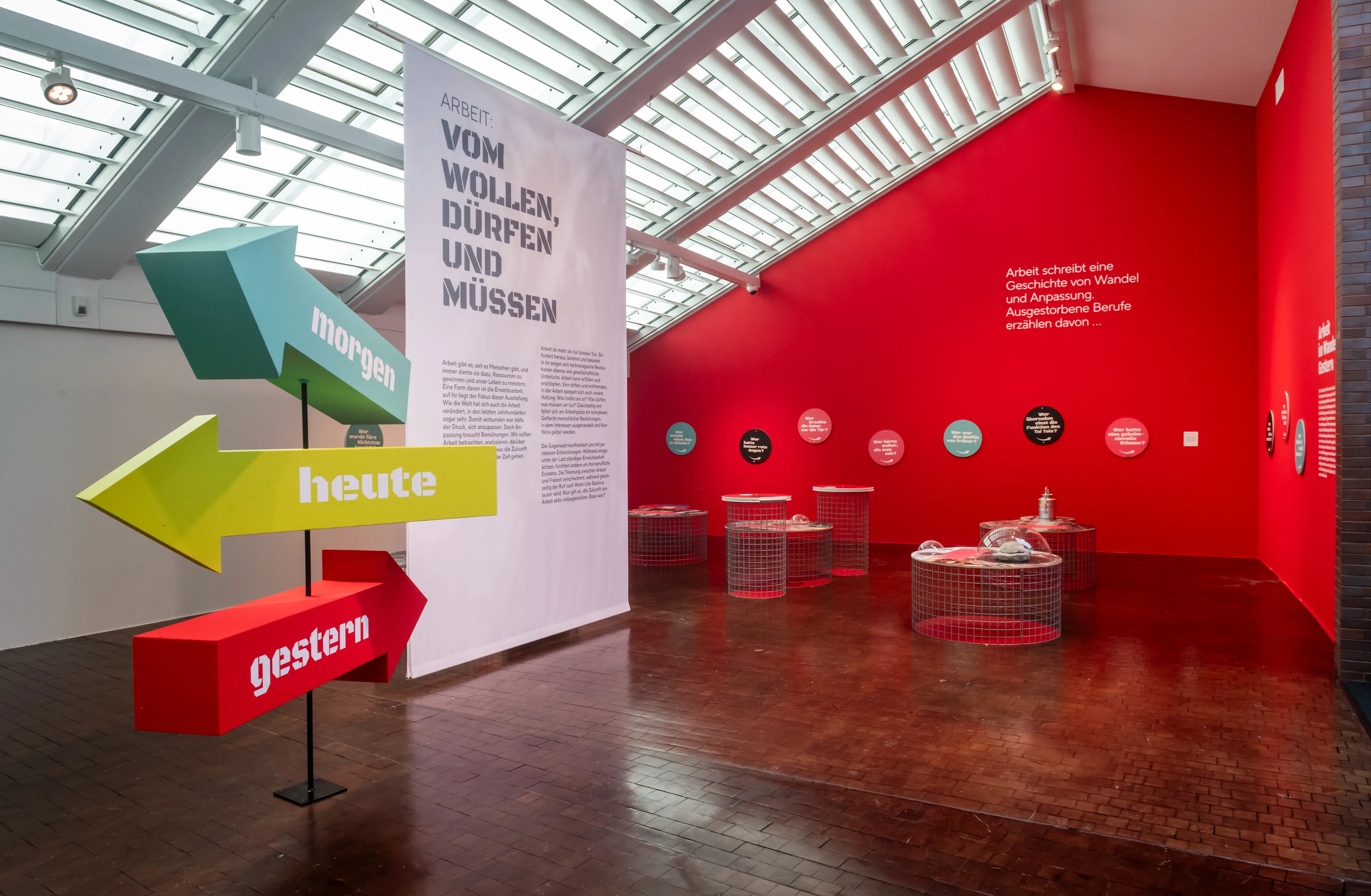 An exhibition room with a red wall, text in German, display tables with objects, and a colorful arrow sign pointing to morgen, heute, and gestern. Skylights illuminate the modern, open space.