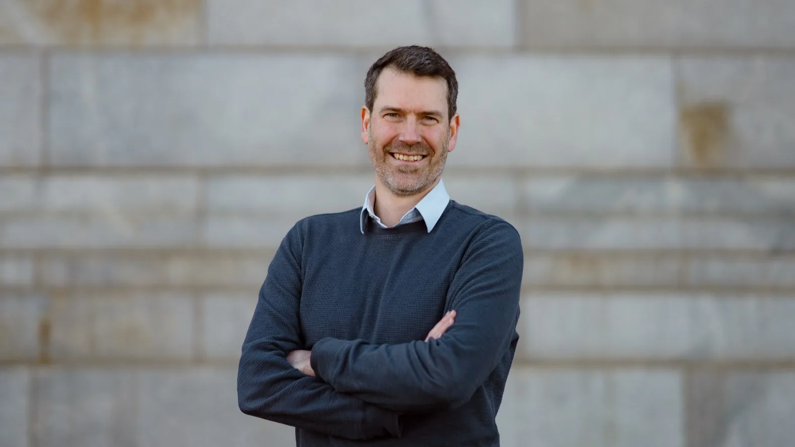 A man with short brown hair and a beard, wearing a dark sweater over a collared shirt, stands smiling with arms crossed in front of a stone wall.
