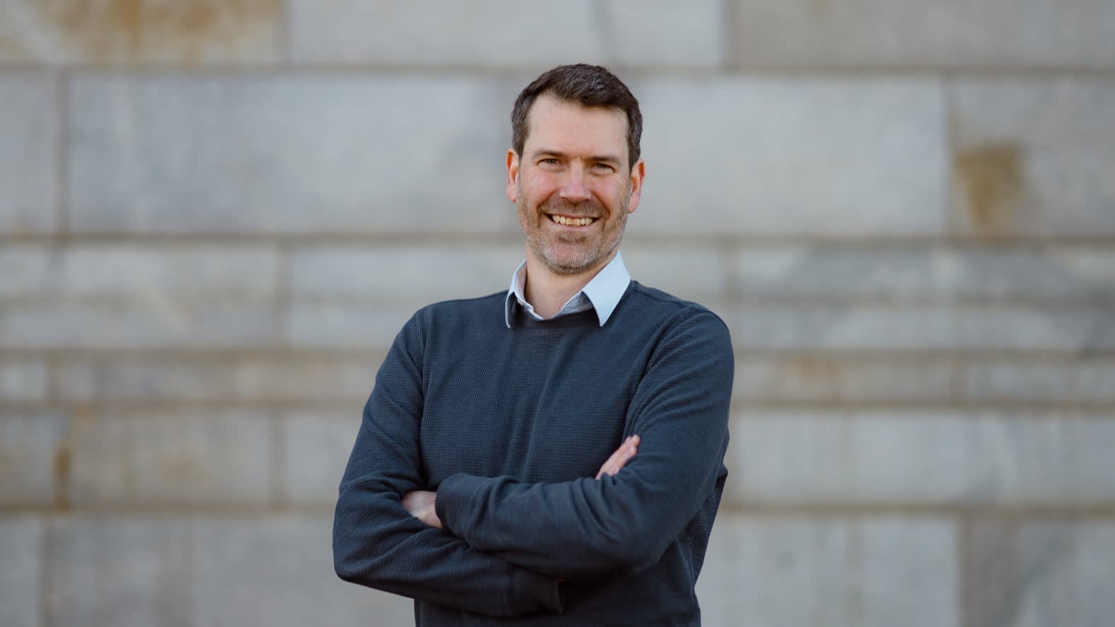 A man with short brown hair and a beard, wearing a dark sweater over a collared shirt, stands smiling with arms crossed in front of a stone wall.