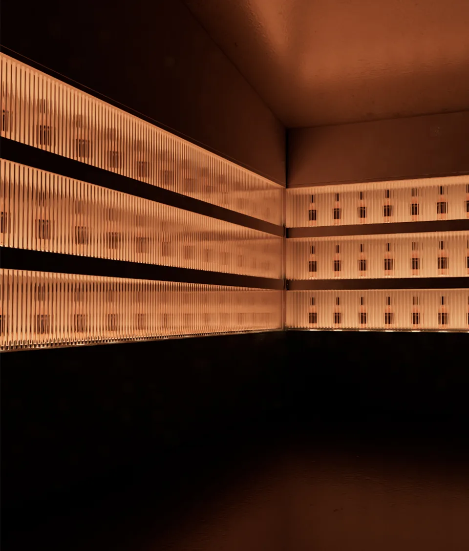 A dimly lit room with three shelves lined with backlit bottles behind vertical slats, creating a warm amber glow and a modern, minimalist ambiance.