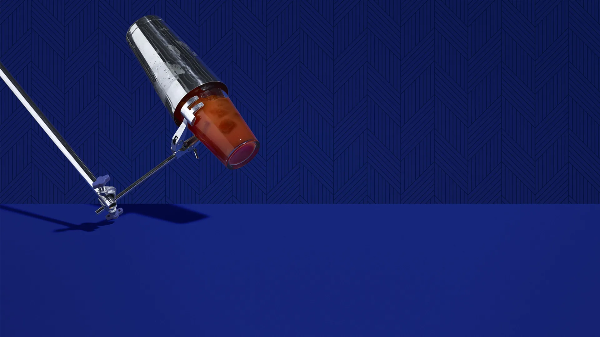 A metal cocktail shaker pours an orange drink onto a blue surface, with a deep blue geometric background and a metal rod supporting the shaker.