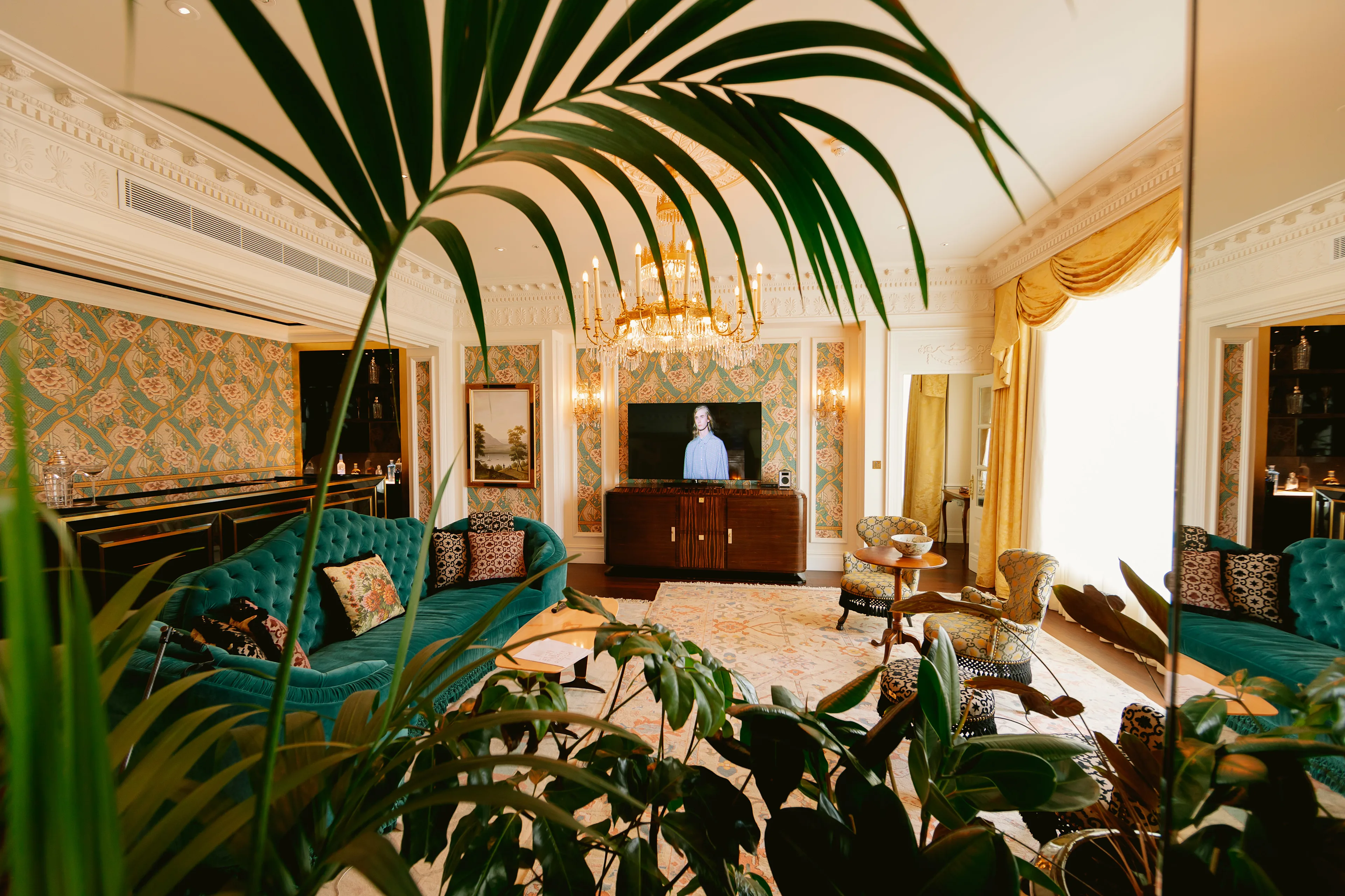 Elegant living room with green velvet sofas, patterned wallpaper, and a chandelier. Lush plants frame the foreground, and a TV with a person on screen is centered on a wooden cabinet beneath large windows with gold curtains.