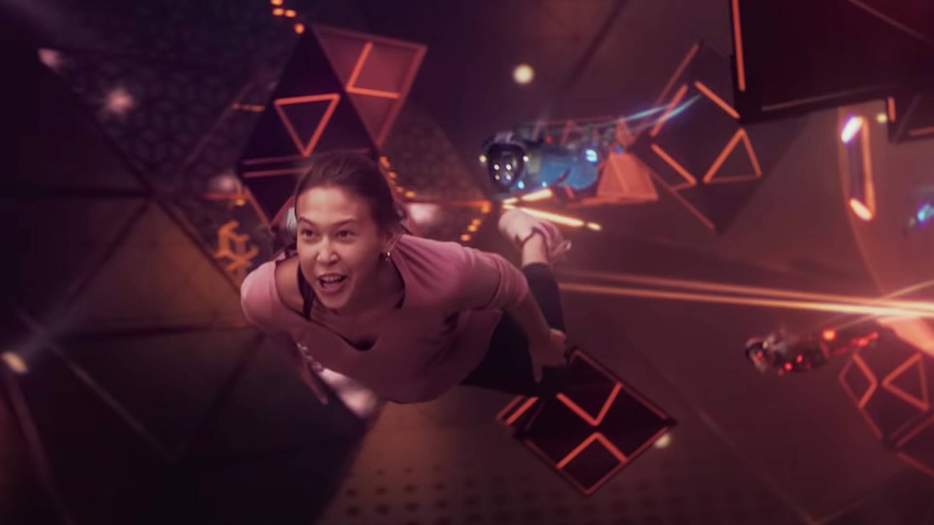 A woman in a pink shirt floats and smiles inside a futuristic, neon-lit virtual environment with geometric shapes and small hovering robots around her.