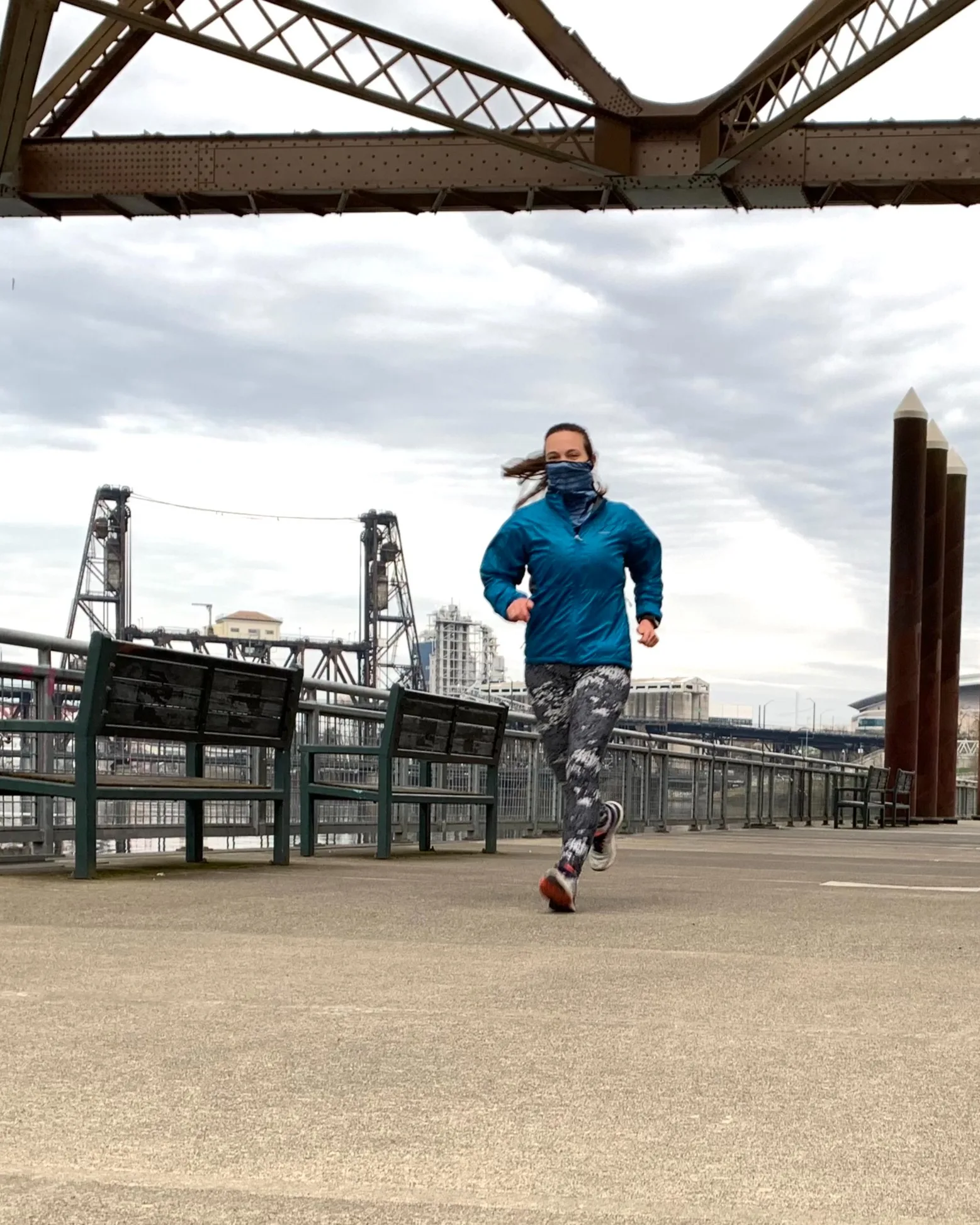 A person wearing a blue jacket and patterned leggings runs on a paved riverside path under a bridge, with city buildings and a steel bridge visible in the background.
