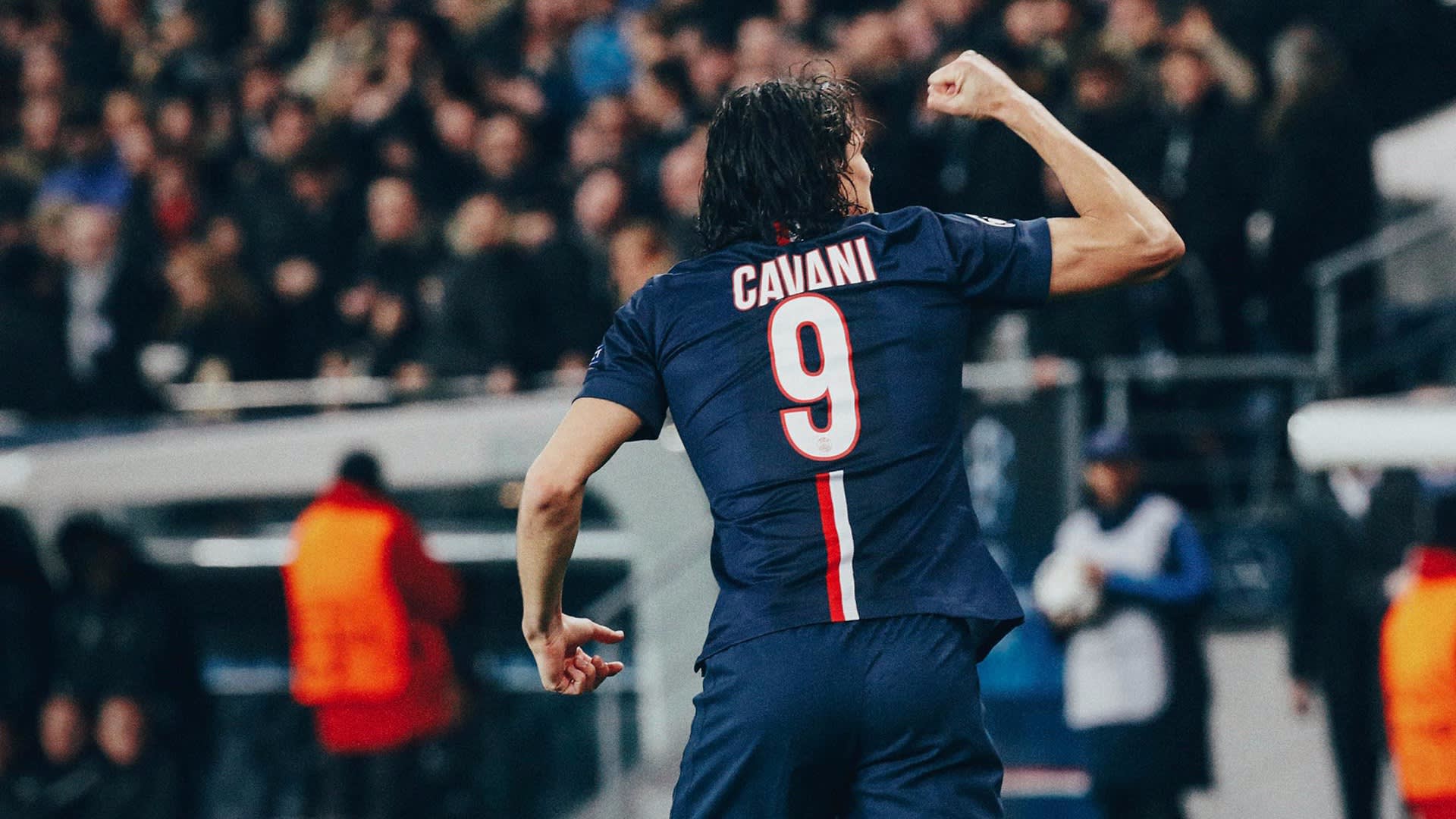 A soccer player wearing a blue jersey with the name Cavani and number 9 raises his fist in celebration on the field, with a blurred crowd in the background.