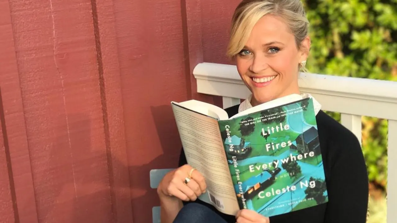 A smiling woman sits on a porch, holding and reading the book Little Fires Everywhere by Celeste Ng. She wears a black top and has blonde hair pulled back. A red wall and green foliage are in the background.