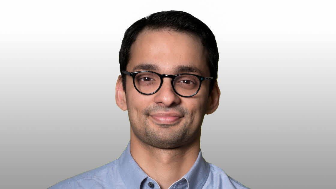 A man with short dark hair and glasses, wearing a light blue collared shirt, smiles slightly against a plain, light gray background.