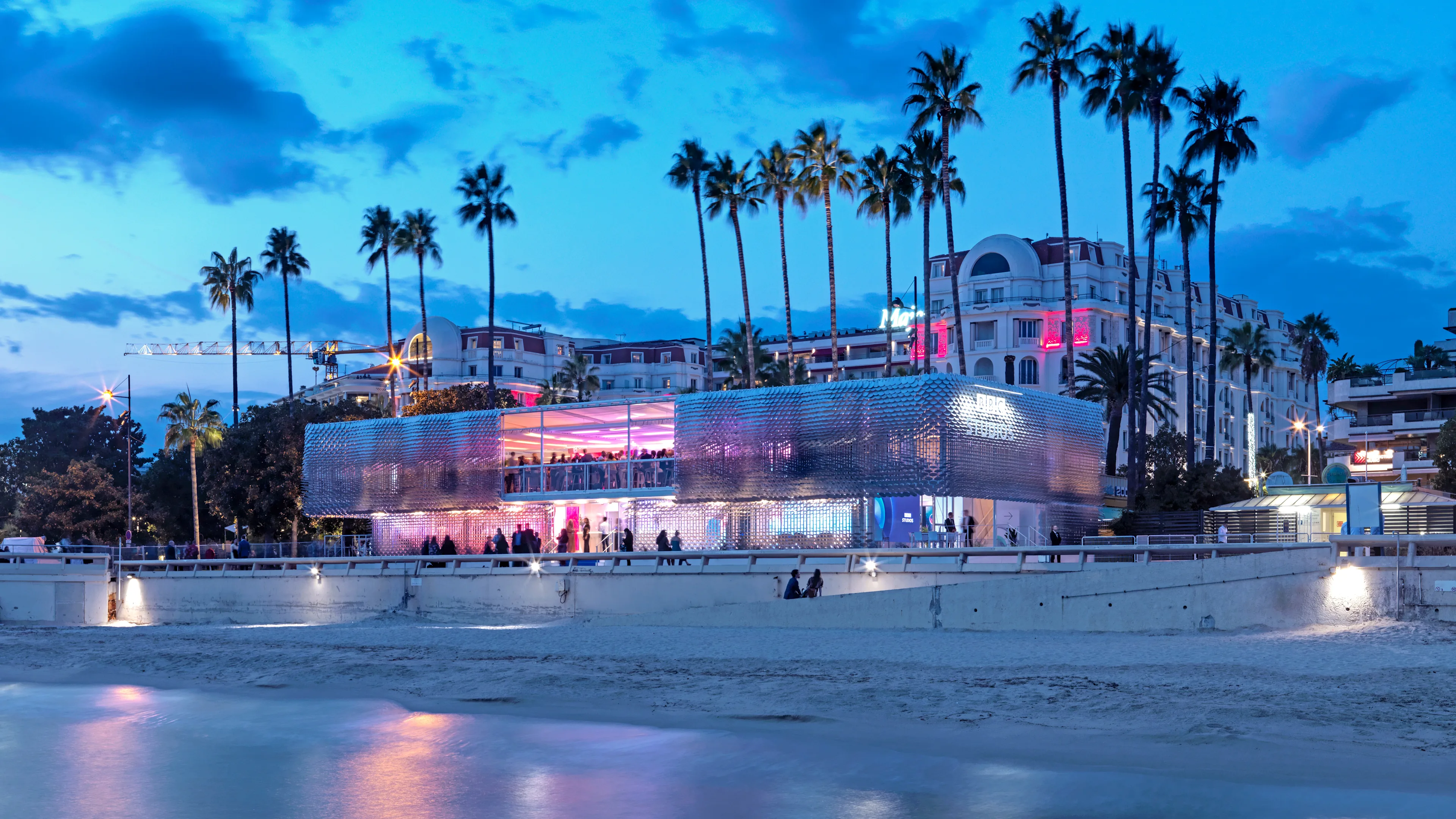 A modern, glass-walled building glows with purple and blue lights near a sandy beach at dusk. Tall palm trees and elegant hotels with illuminated windows are in the background. People gather along the water’s edge.
