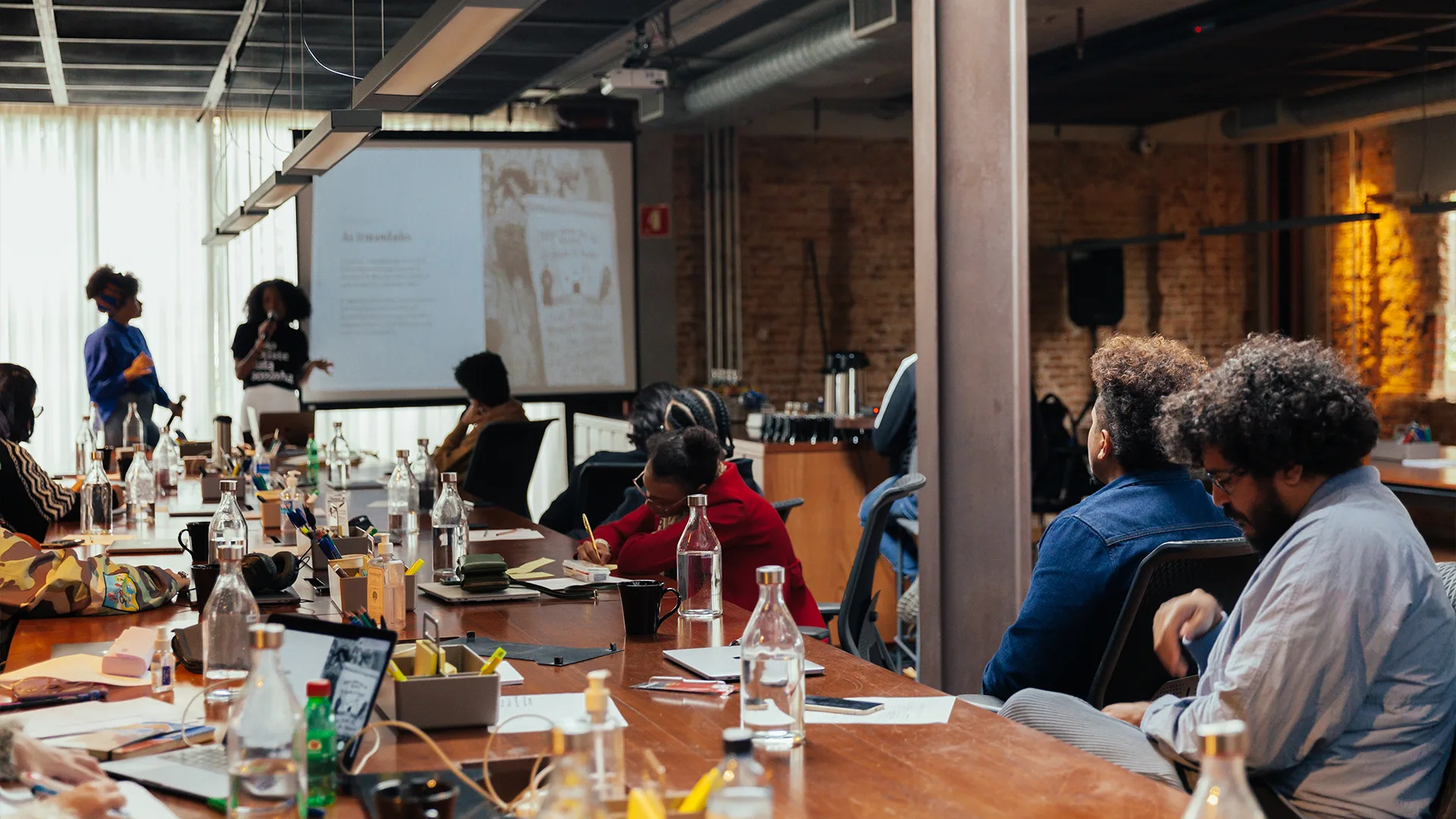People sit around a large conference table covered with bottles, papers, and laptops, while two presenters stand by a screen displaying a presentation in a modern, industrial-style room.