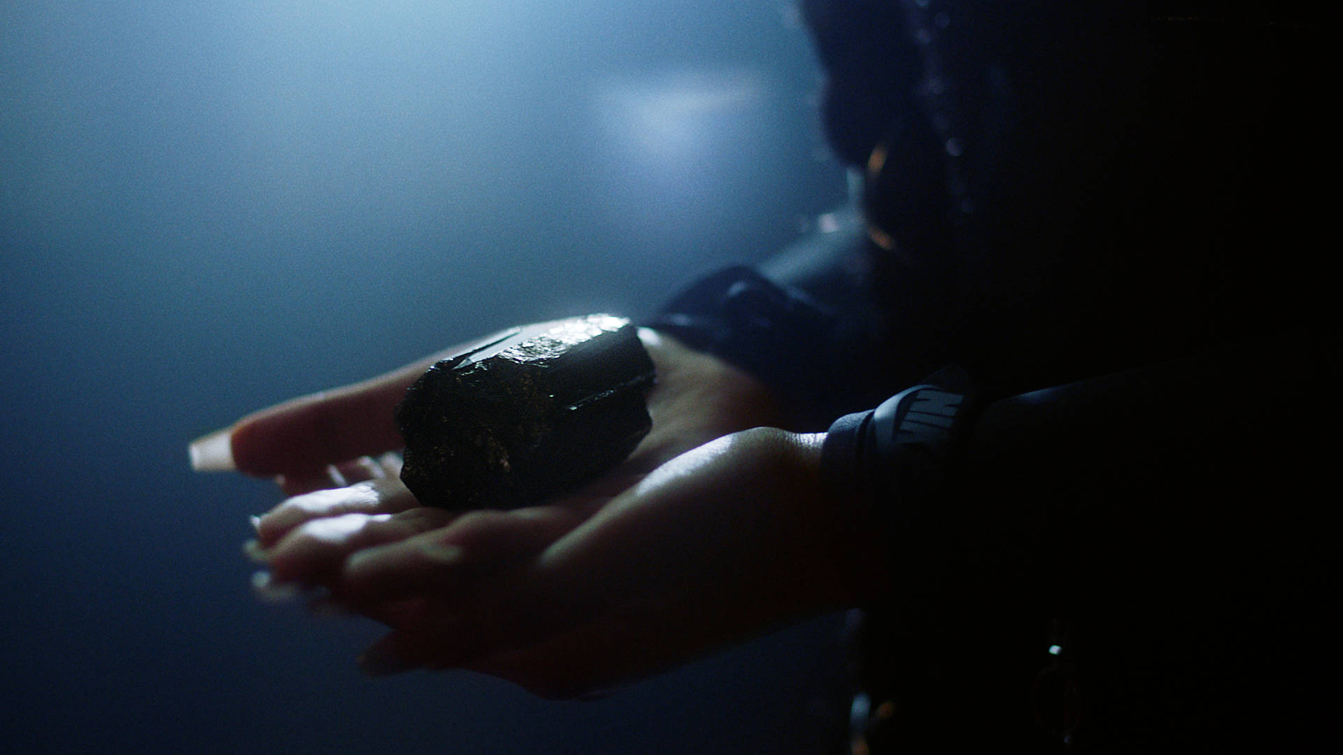 A person with long nails holds a shiny, dark rock in their hands under moody, blue-toned lighting. The image is close-up, focusing on the hands and the object, with the background blurred.